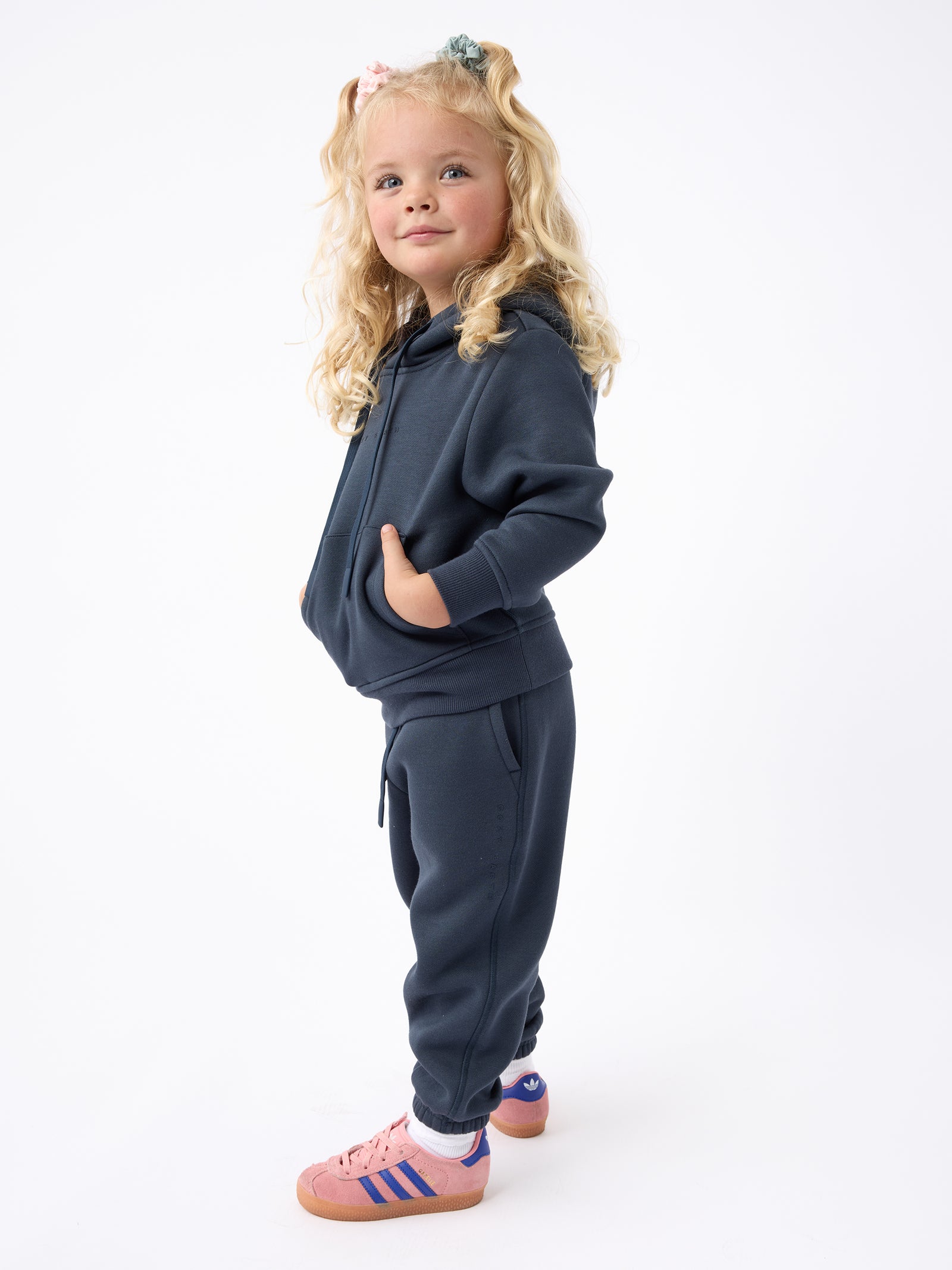 A young child with long blonde hair stands confidently, dressed in a Cozy Earth Kid's CityScape Hoodie and sweatpants. With one hand tucked in a pocket, they sport pink sneakers with blue stripes against a plain white backdrop. 