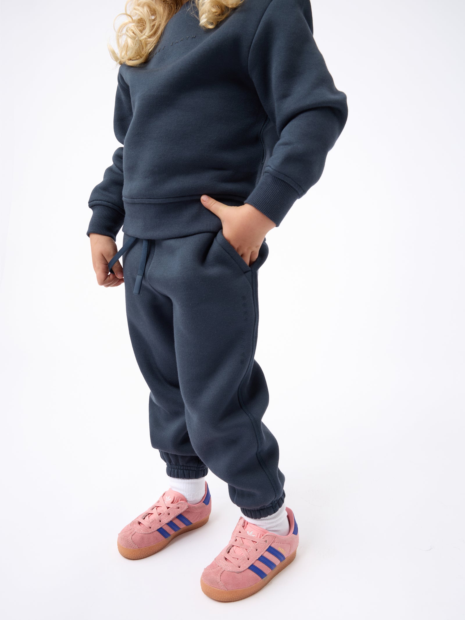 A child wearing a dark blue Kid's CityScape Sweatpant from Cozy Earth, with one hand in the pocket. They have long blonde hair and are also wearing pink Adidas sneakers with blue stripes and white socks, standing against a plain white background. 