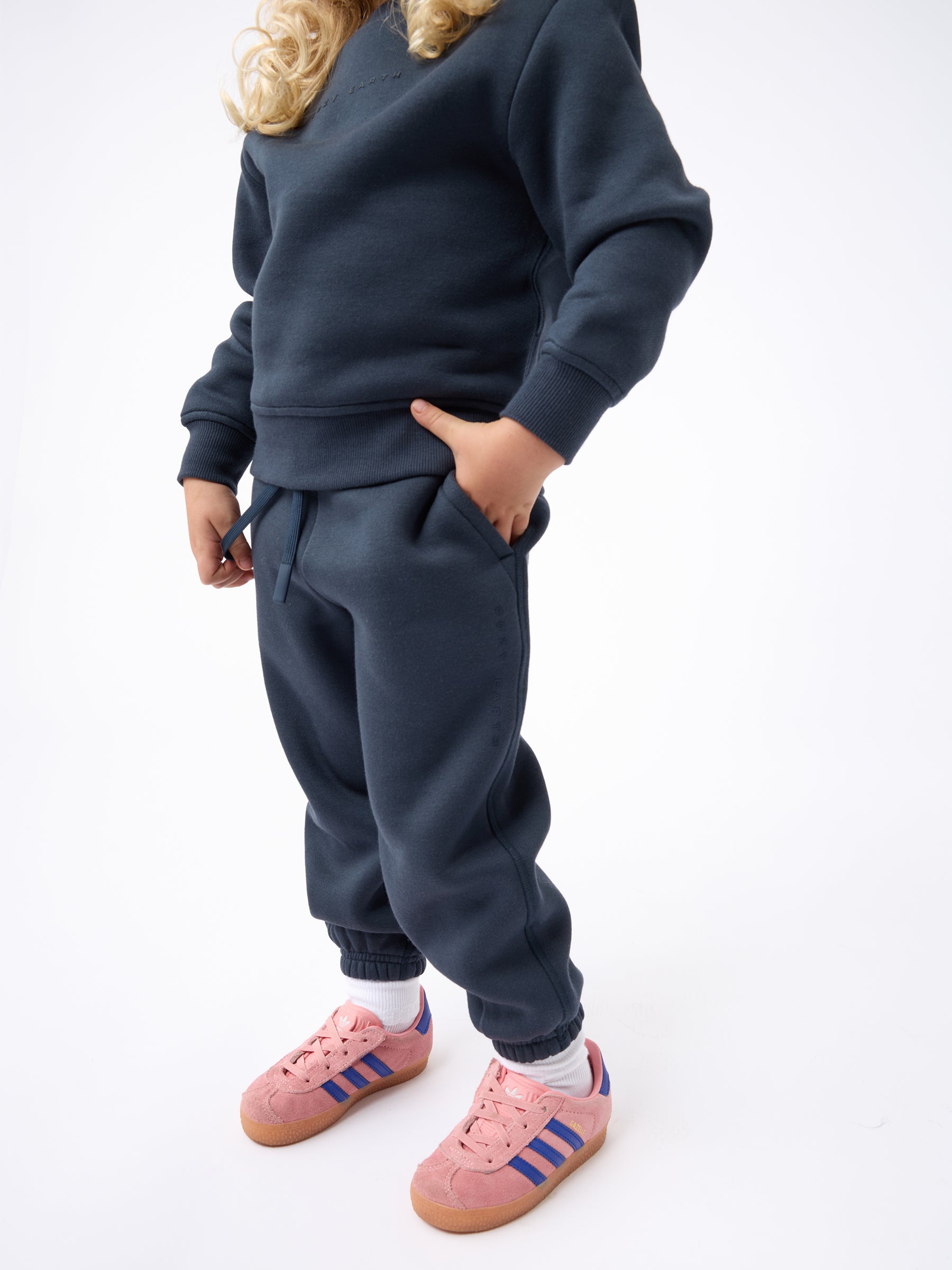 A child wearing a dark blue Kid's CityScape Sweatpant from Cozy Earth, with one hand in the pocket. They have long blonde hair and are also wearing pink Adidas sneakers with blue stripes and white socks, standing against a plain white background. |Color:Eclipse