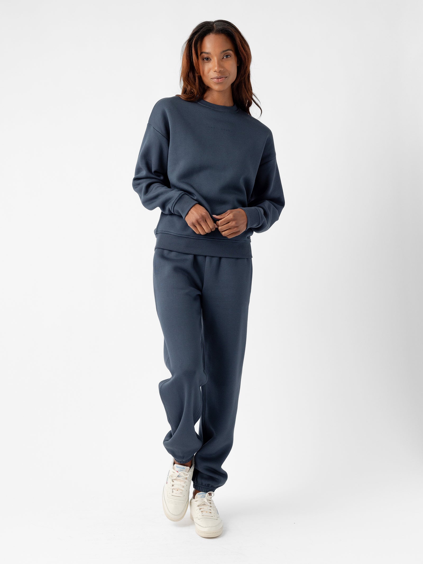 A person stands against a plain white background. They have long hair and are wearing Cozy Earth's Women's CityScape Crewneck in navy blue, paired with matching sweatpants. They are also wearing white sneakers. The person is looking at the camera with a relaxed expression, with one hand in a pocket. 