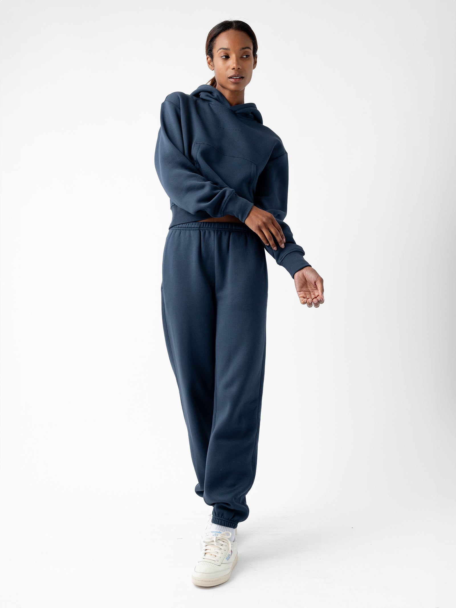 A person stands against a white background, wearing a matching Women's CityScape Cropped Hoodie and sweatpants from Cozy Earth in navy blue. They have white sneakers on, with one hand raised slightly in front of their body while the other hangs by their side. 