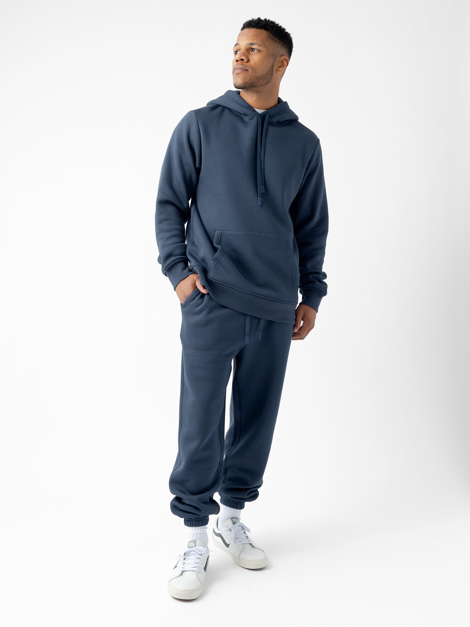 A man stands against a plain white background wearing a matching Men's CityScape Hoodie and sweatpants set from Cozy Earth. He has his hands in his hoodie pockets and is looking to his left. He is also wearing white sneakers. 