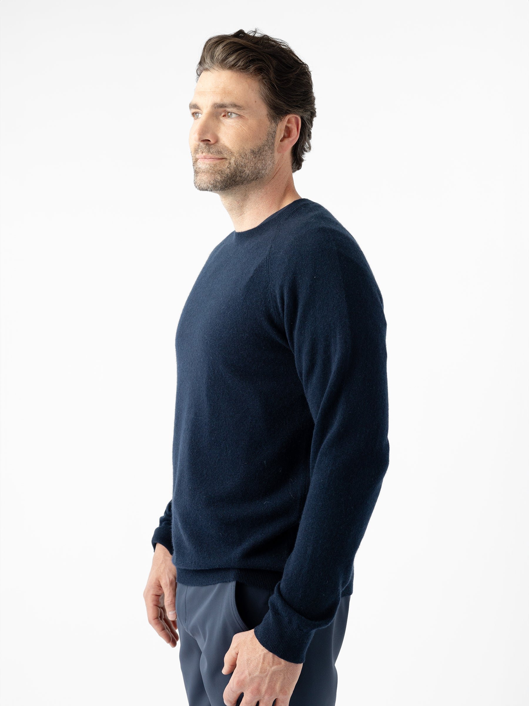A man with short, dark hair and a beard is standing sideways, facing forward. He is wearing the Men's Crewneck Sweater by Cozy Earth in a dark navy color, paired with gray pants. The background is entirely white, giving a clean and minimalist look to the image. 