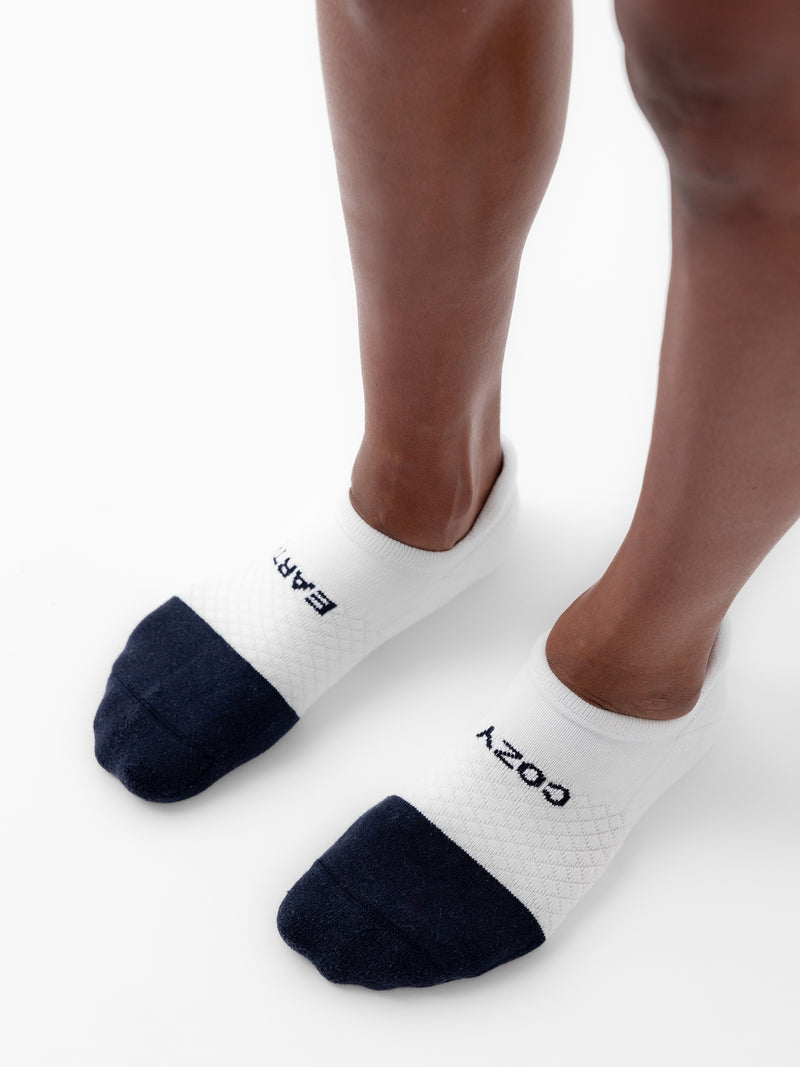 Close-up of someone wearing white ankle socks with black toes from the Essential Ankle Sock 2-Pack by Cozy Earth. The socks have the words "EARTH" and "COZY" written on them in black, with one word on each sock. The person is standing on a white surface.