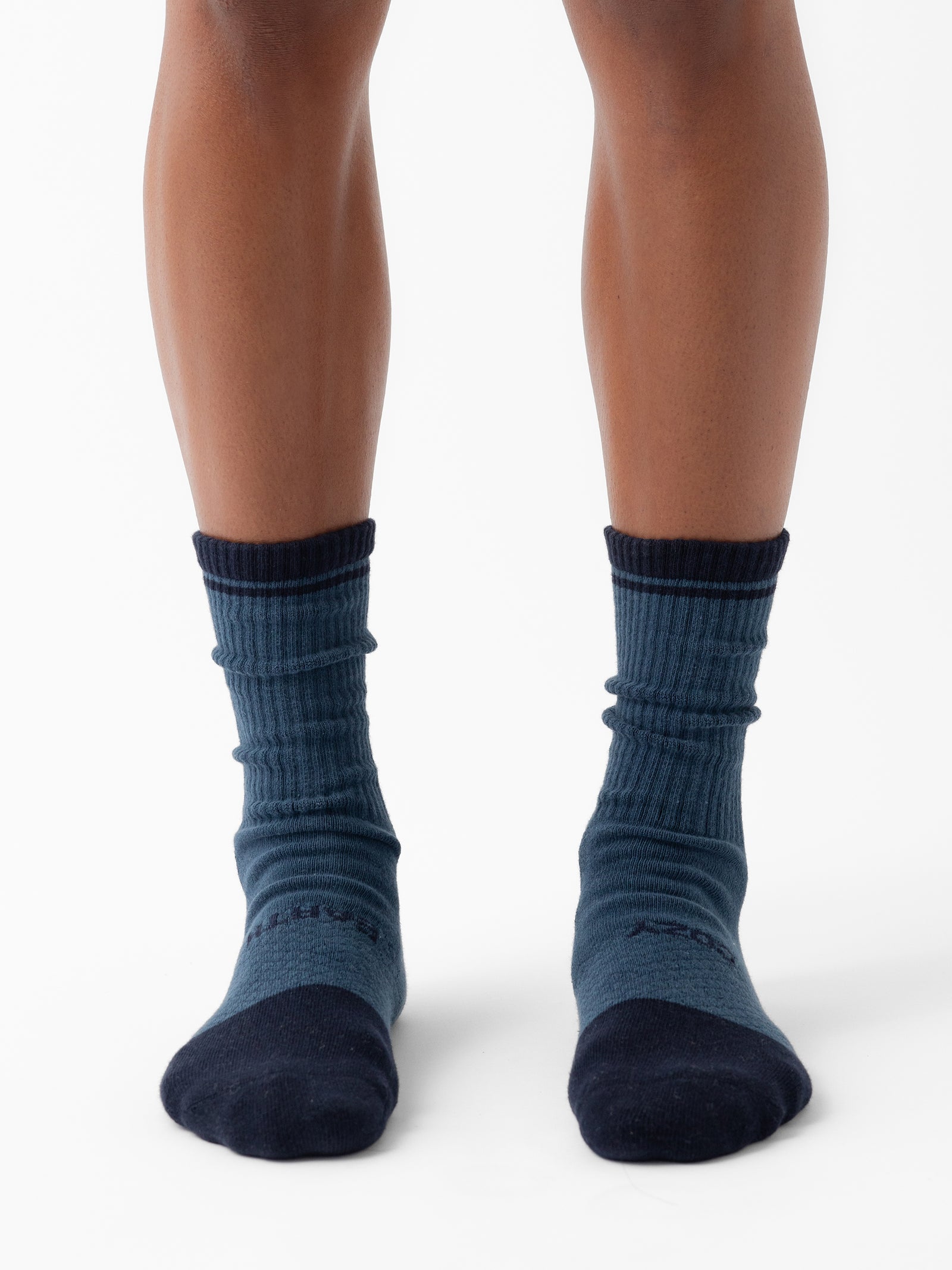 A person stands facing forward wearing Cozy Earth's Essential Calf Sock 2-Pack in dark blue, featuring black toes and heels. The person's lower legs are visible against a plain white background, with the socks appearing slightly wrinkled around the ankles. 