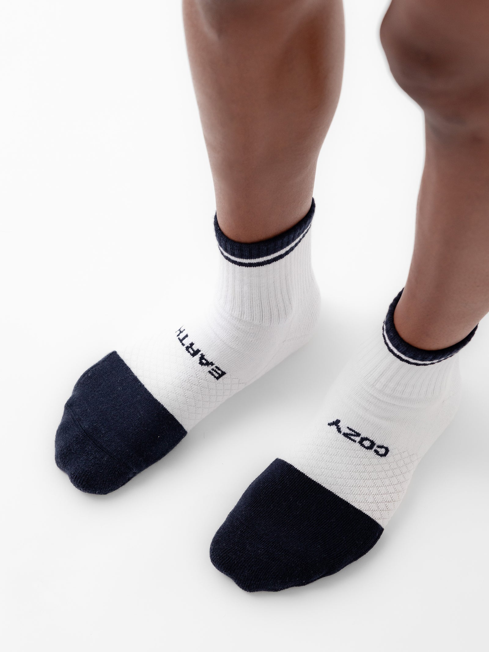 A close-up image of a person's feet wearing a pair of Cozy Earth's Essential Quarter Socks from the 2-Pack collection, featuring white socks with black toes, heels, and cuffs. The left sock is emblazoned with "EARTH," while the right one displays "COZY." The person is standing on a light background. 