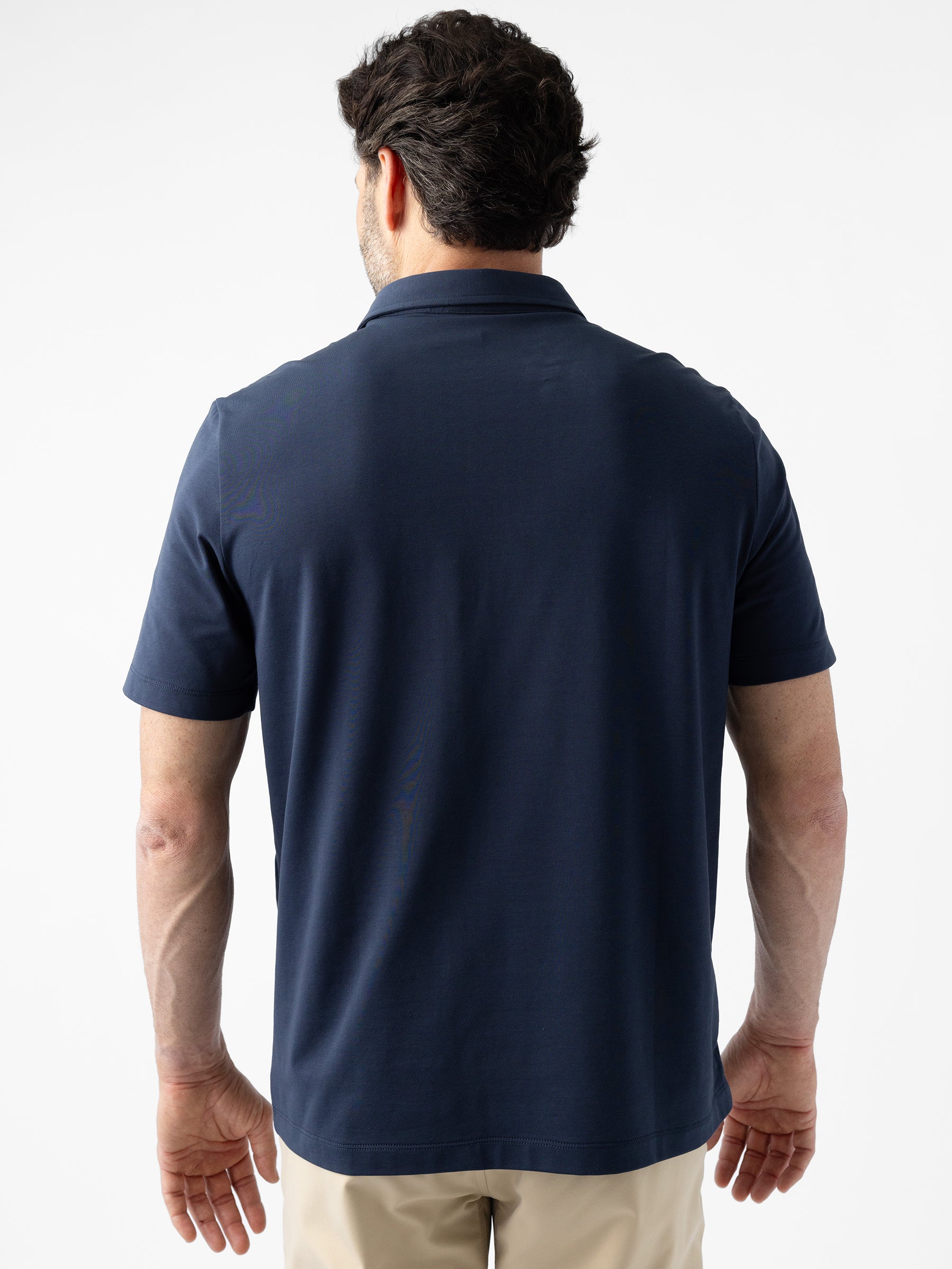 A man with short dark hair faces away while wearing Cozy Earth's Men's Everyday Polo in navy blue and beige pants, standing against a plain white background. |Color:Eclipse