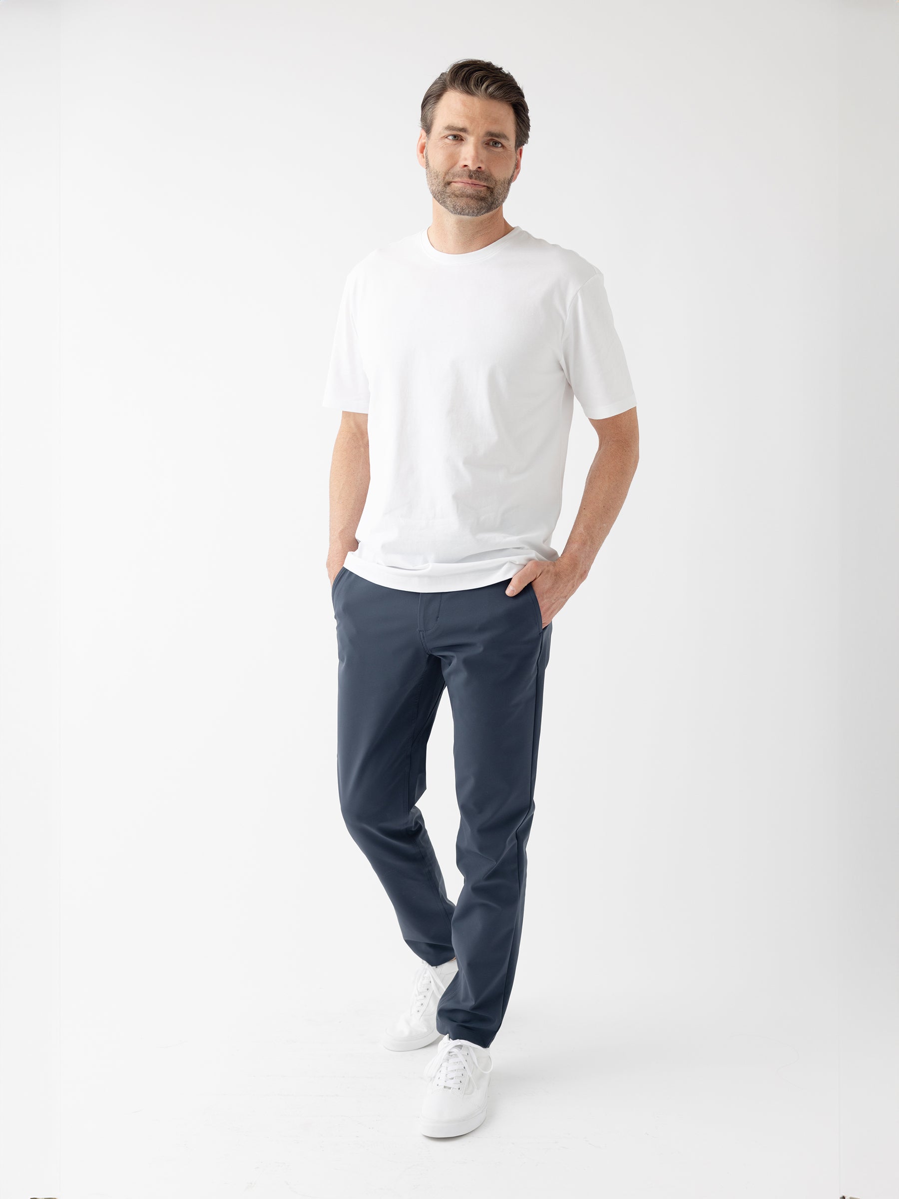 A man stands confidently against a white background. He is wearing a plain white T-shirt, Cozy Earth's Men's Everywhere Pant 30L in dark blue, and white sneakers. His hands are casually tucked into his pants pockets. He has short hair and a neatly trimmed beard. |Color:Eclipse