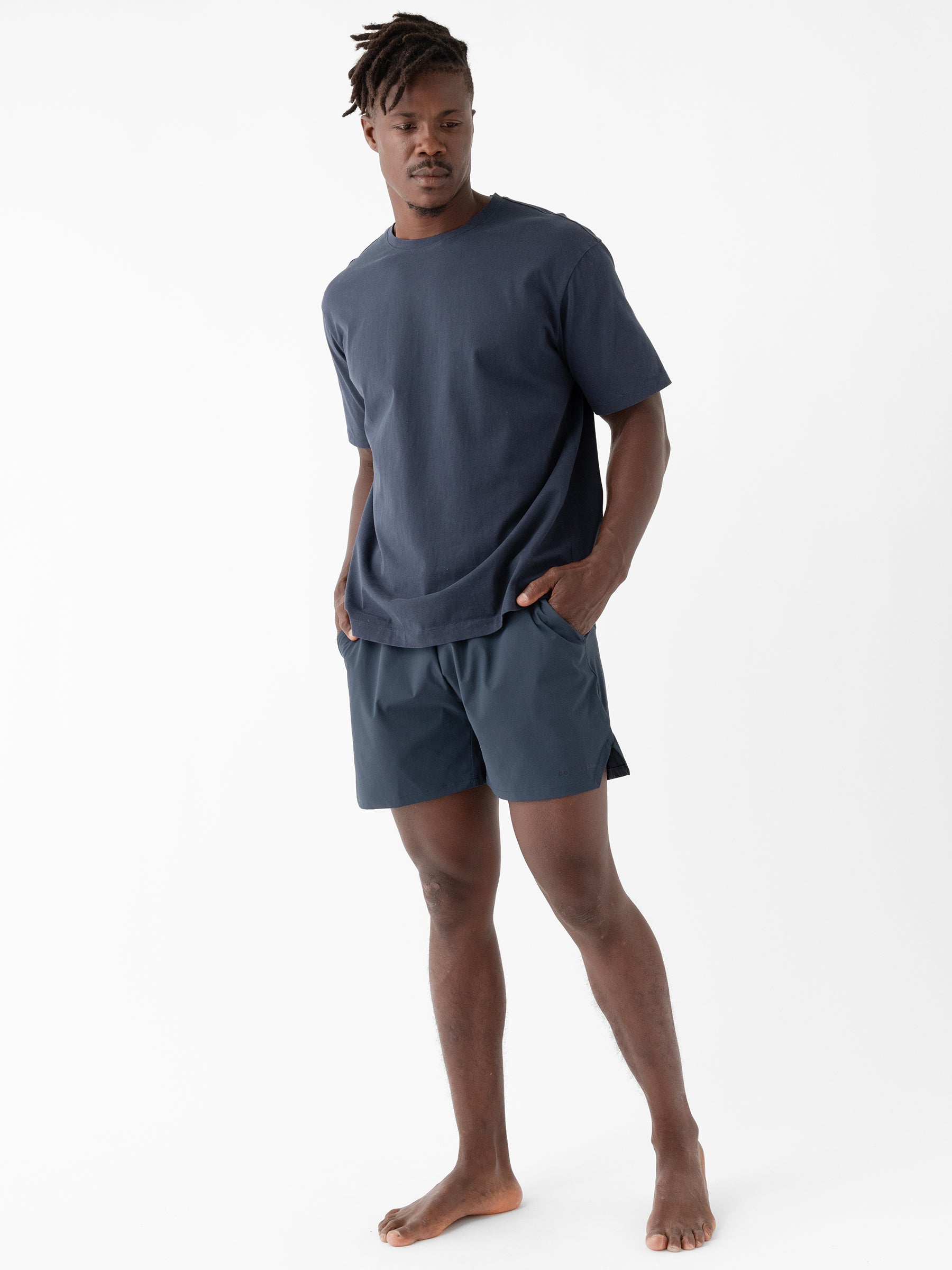 A man stands barefoot against a plain white background, wearing a loose-fitting navy blue T-shirt and Cozy Earth's Men's Performance Sleep Short. His hands are in his pockets, and his head is slightly tilted as he looks downward. He has short dreadlocks and a relaxed, casual posture. |Color:Eclipse