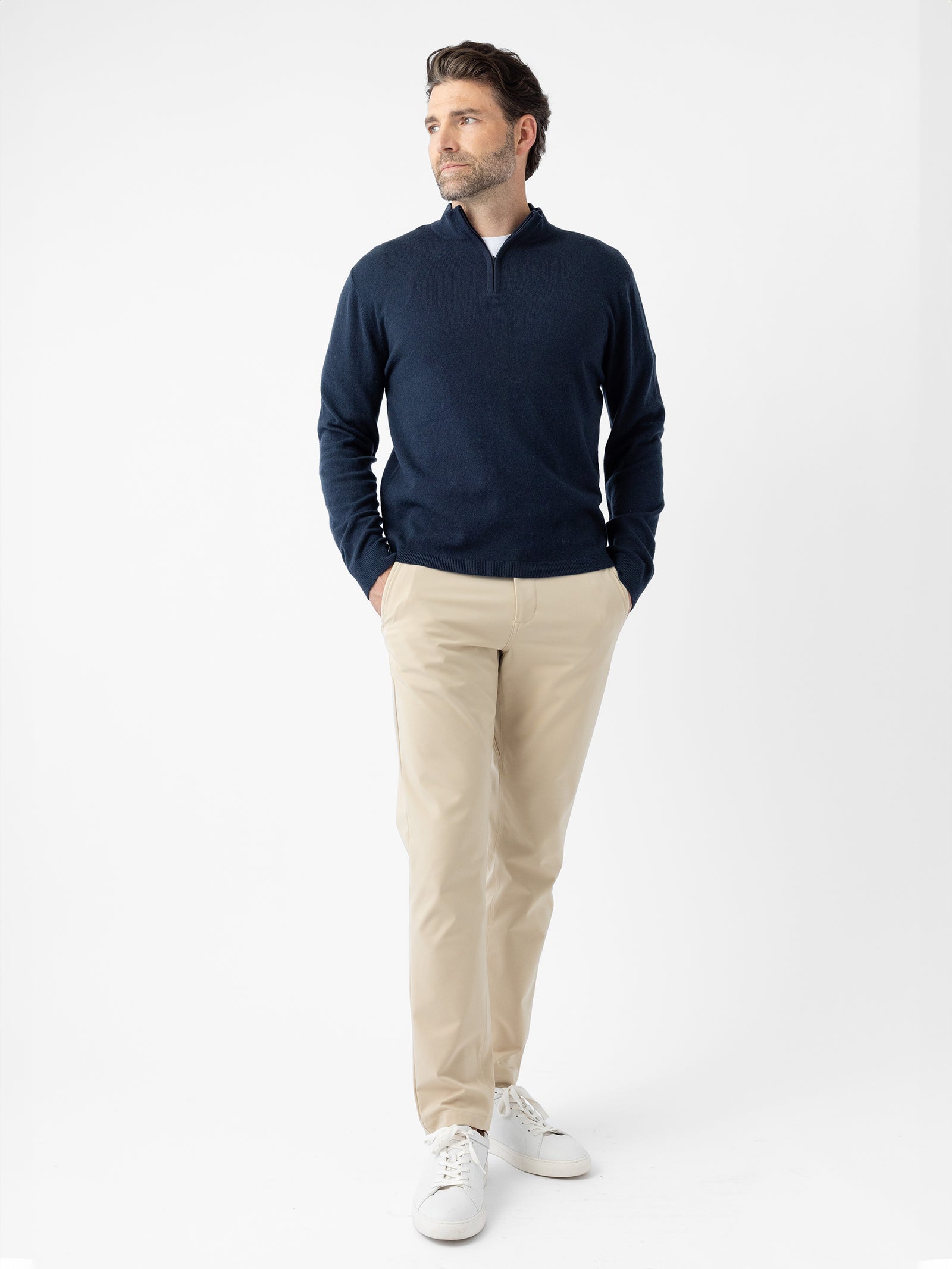 A man stands against a plain white background wearing Cozy Earth's Men's Quarter Zip Sweater in dark blue, along with beige pants and white sneakers. His hands are in his pockets as he gazes slightly to his right. 