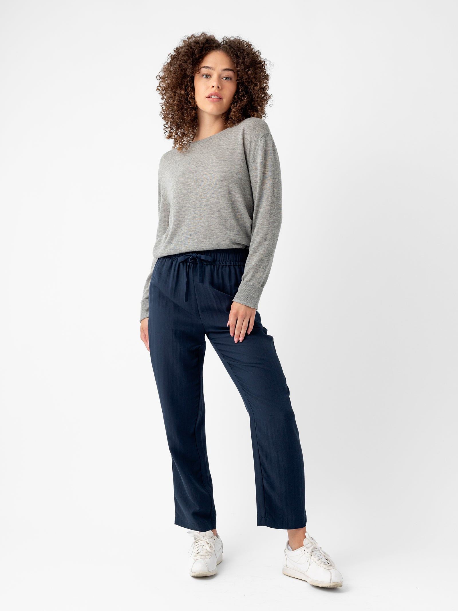 A woman with curly hair is standing against a plain white background. She is wearing a light grey long-sleeve shirt, Cozy Earth’s dark blue Women's Sunset Cropped Pant, and white sneakers. She has a relaxed pose with one hand in her pocket and a neutral expression on her face. 