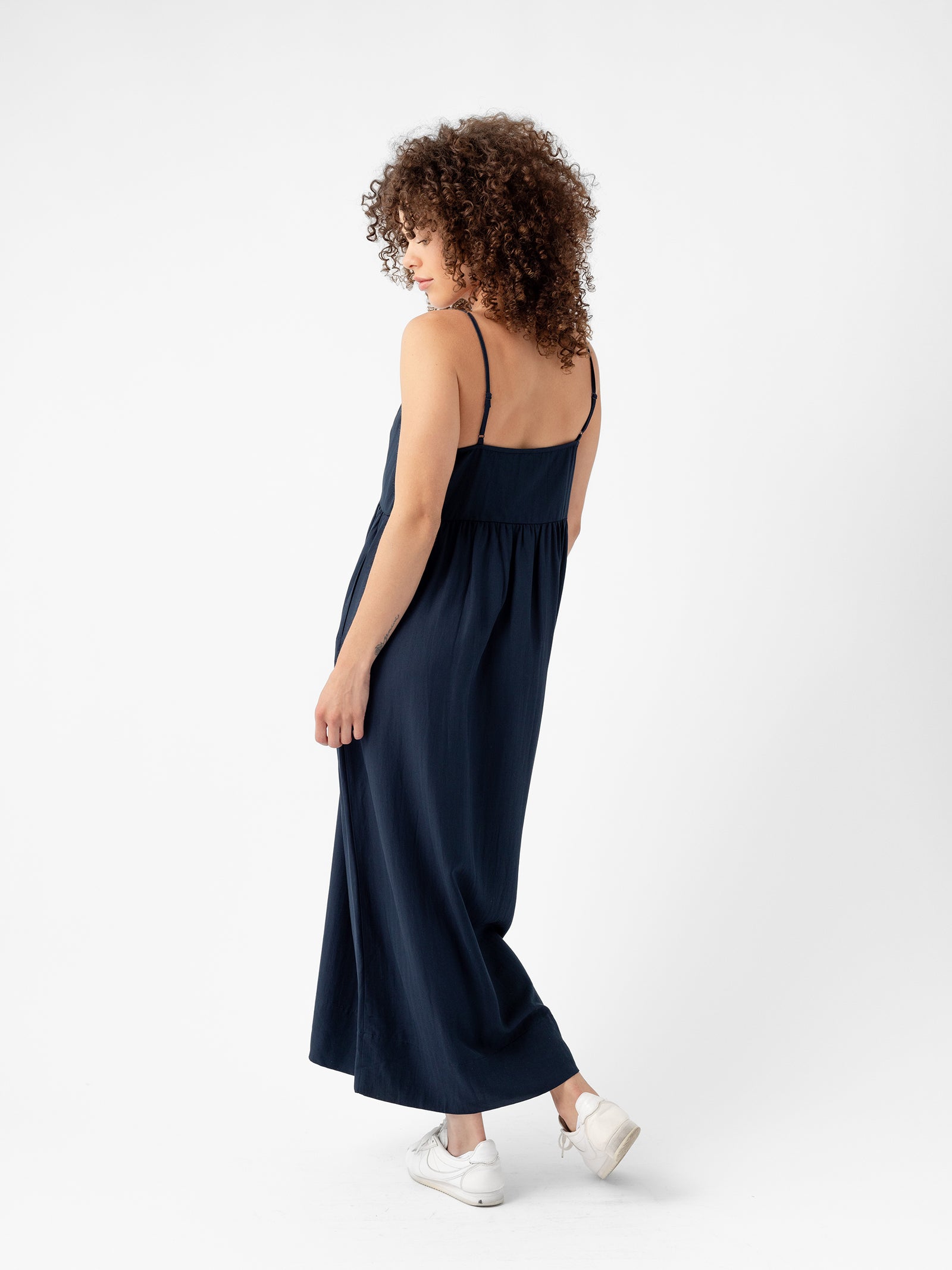 A woman with curly hair is standing against a white background, wearing the Cozy Earth Women's Sunset Gathered Dress in dark blue. The dress features thin straps and is sleeveless. She pairs it with white sneakers and slightly turns to the side, looking down with her left hand resting on her thigh. 