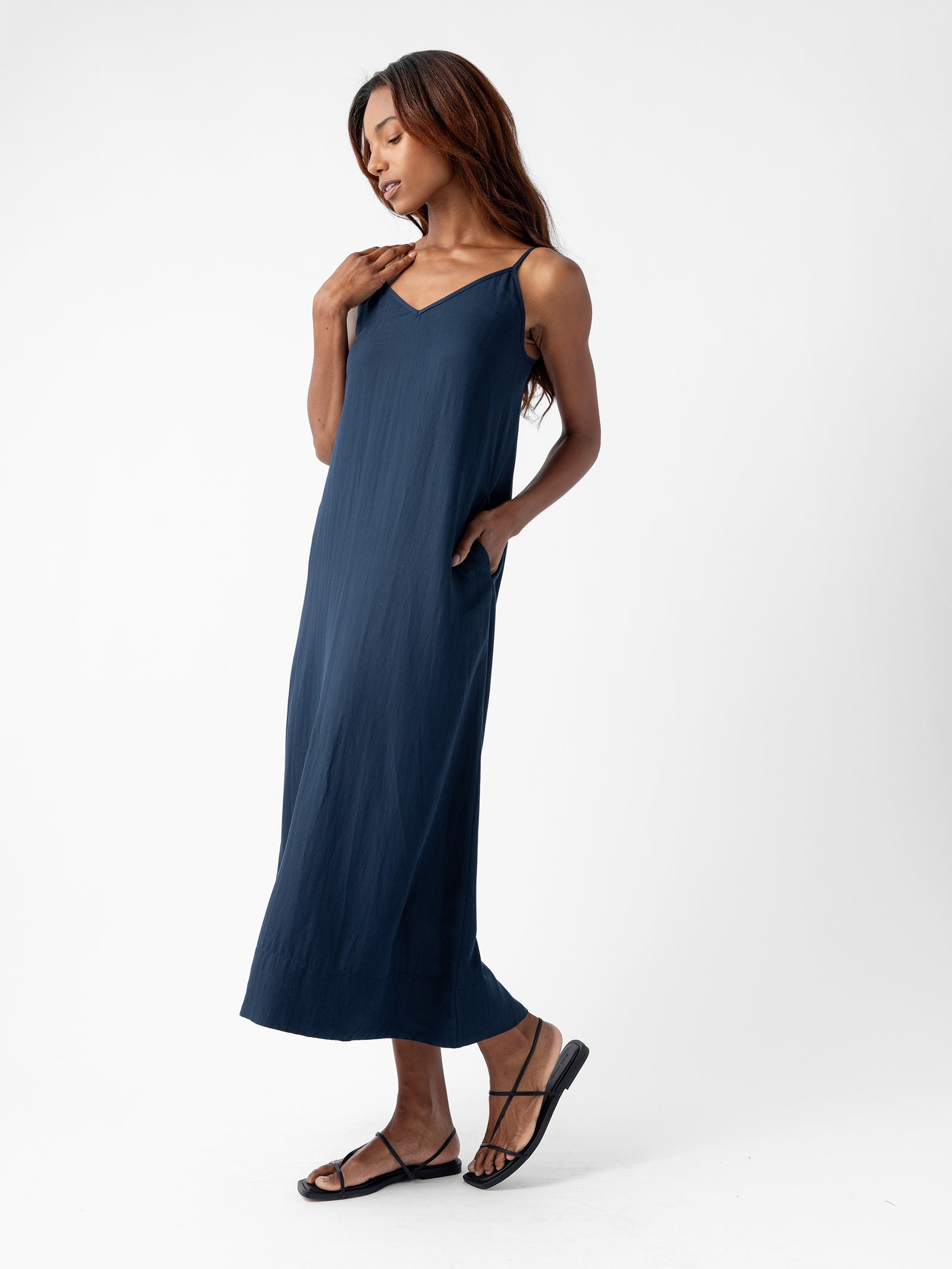 A woman with long hair is wearing the Women's Sunset Slip Dress by Cozy Earth, which features a navy blue, sleeveless, ankle-length design with a V-neckline. She is posing with one hand touching her neck and the other hand in the dress pocket while also wearing black sandals. The background is plain white. 