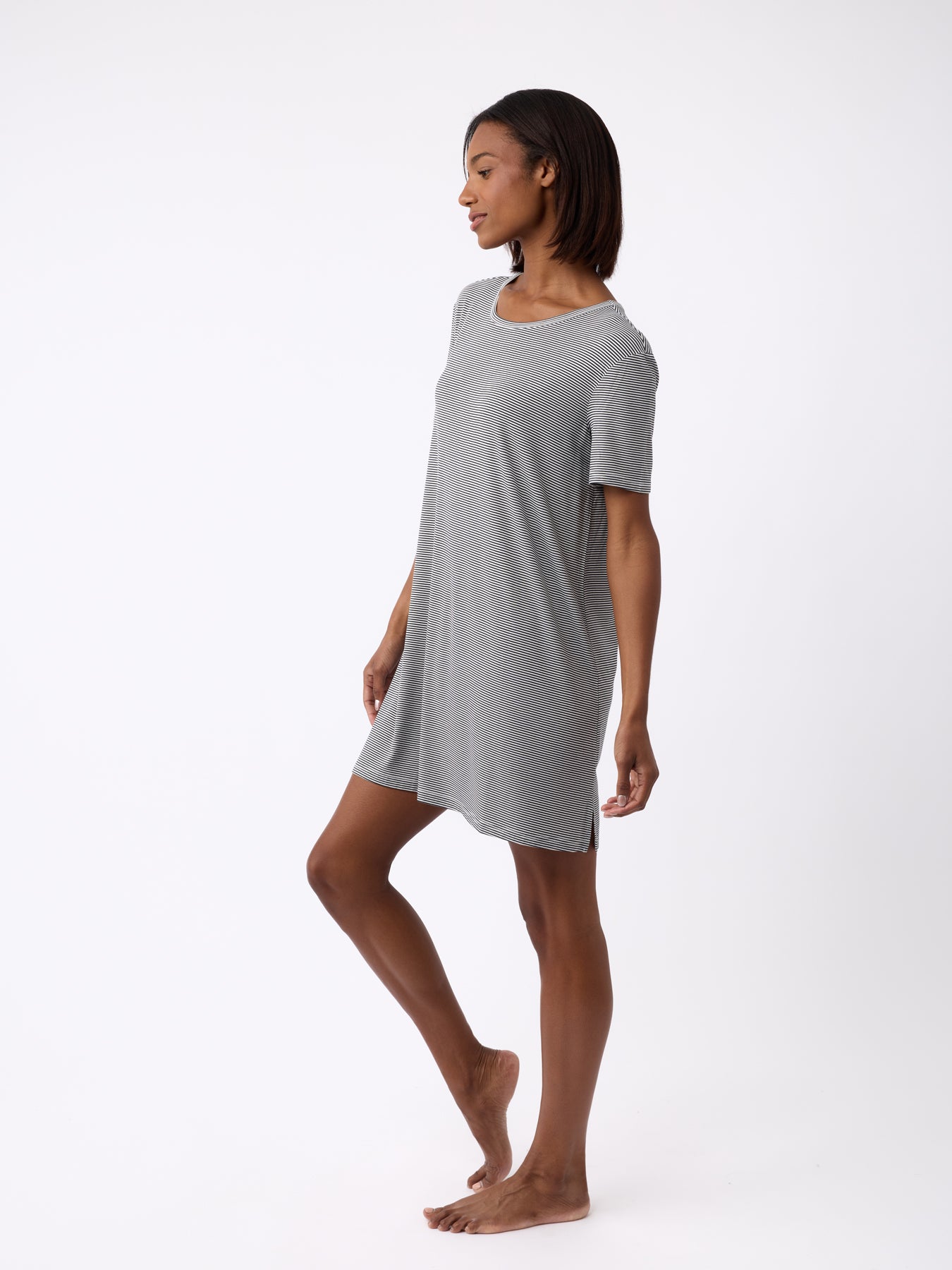 A woman stands barefoot against a white background wearing the Cozy Earth Women's Bamboo Stretch-Knit Sleep Dress. She has shoulder-length dark hair and is posed side-facing with a relaxed posture and neutral expression. 