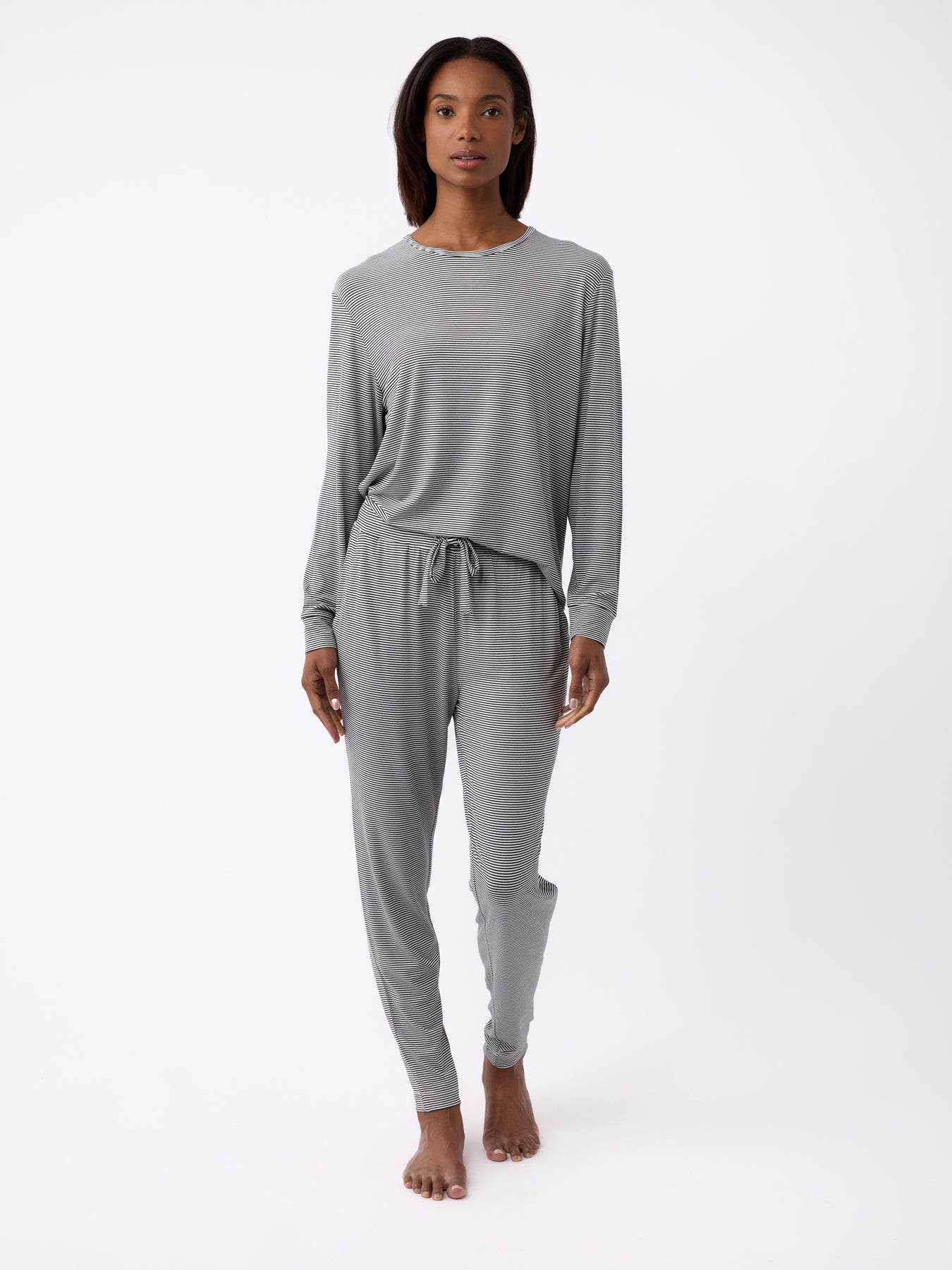 A person stands barefoot against a plain white background, wearing Cozy Earth's Women's Bamboo Stretch-Knit Long Sleeve Lounge Tee in gray with a drawstring waist. 