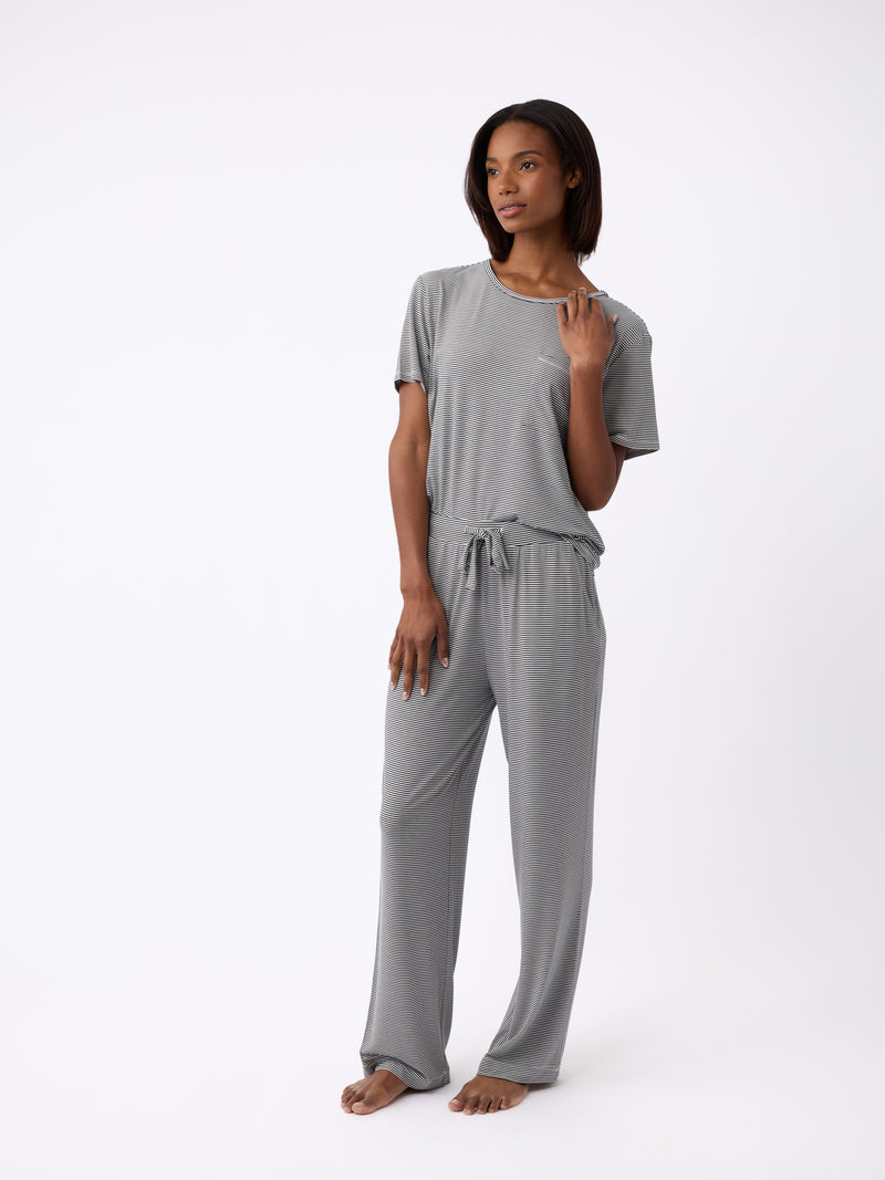 A woman stands barefoot against a white background, her hand raised to her neck. She's wearing Cozy Earth's loose-fitting Women's Bamboo Stretch-Knit Pant in gray, paired with a matching T-shirt.