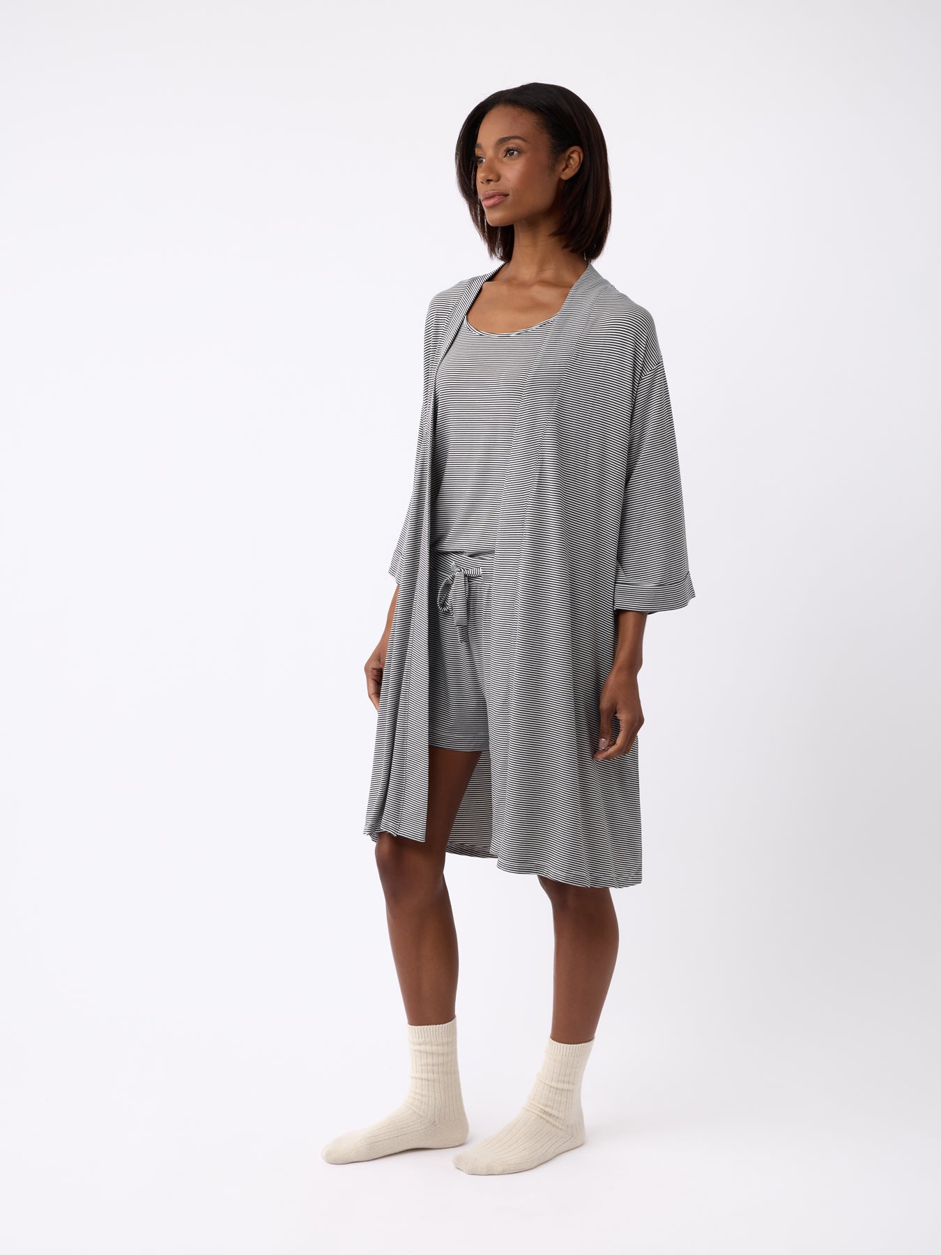 Clad in a Cozy Earth Women's Bamboo Stretch-Knit Kimono Robe over matching shorts and tank top, a person with short dark hair sports cream-colored socks against a plain white backdrop. 