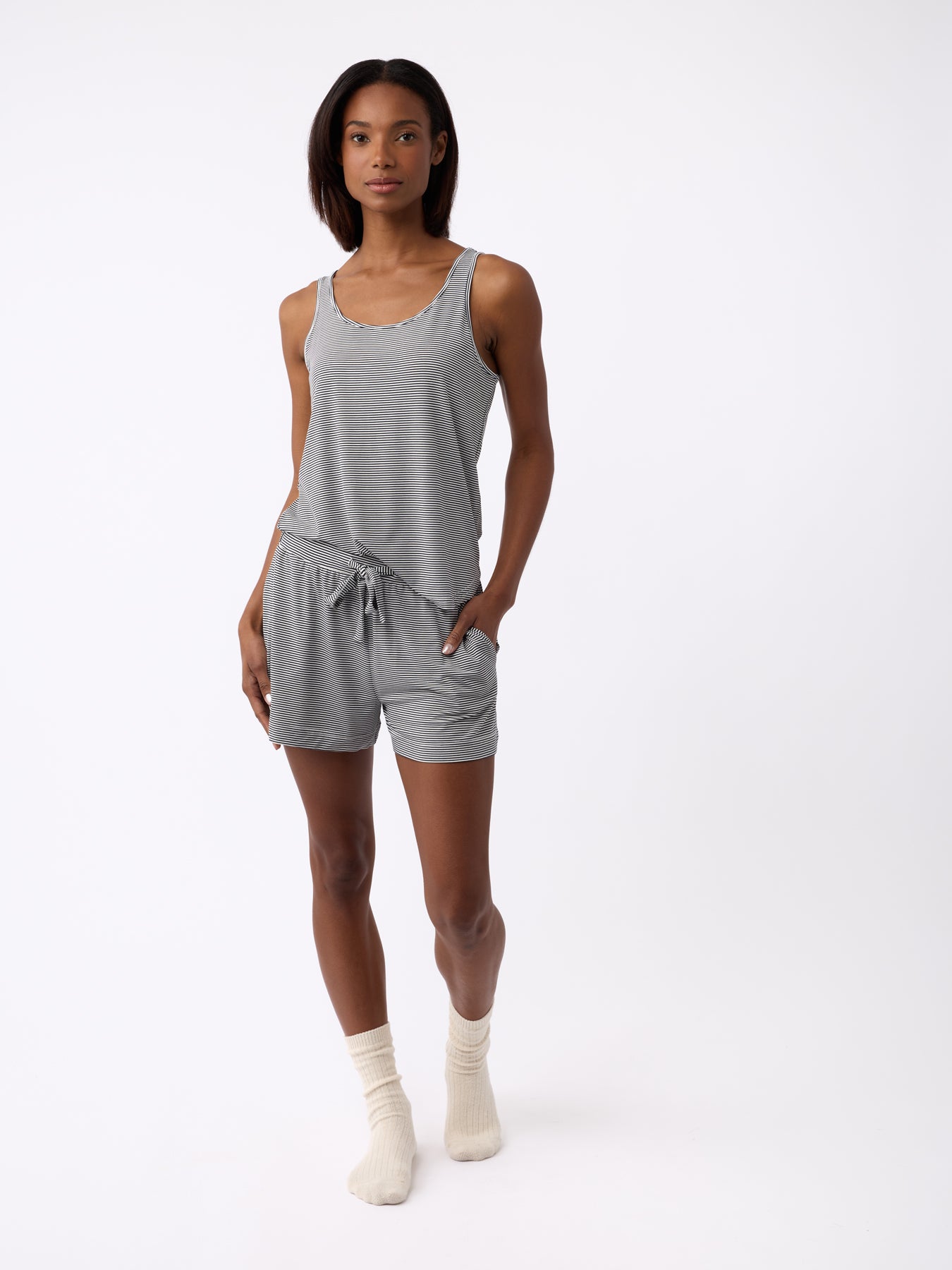 A person stands against a plain background wearing Cozy Earth's Women's Bamboo Stretch-Knit Shorts in gray, paired with a matching tank top and white socks, with one hand in their pocket. 