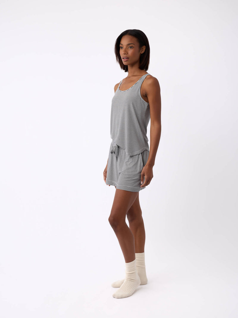 A person with shoulder-length hair stands against a plain background, wearing Cozy Earth's Women's Bamboo Stretch-Knit Lounge Tank in gray and matching shorts. They are also wearing beige socks and facing slightly to the side.