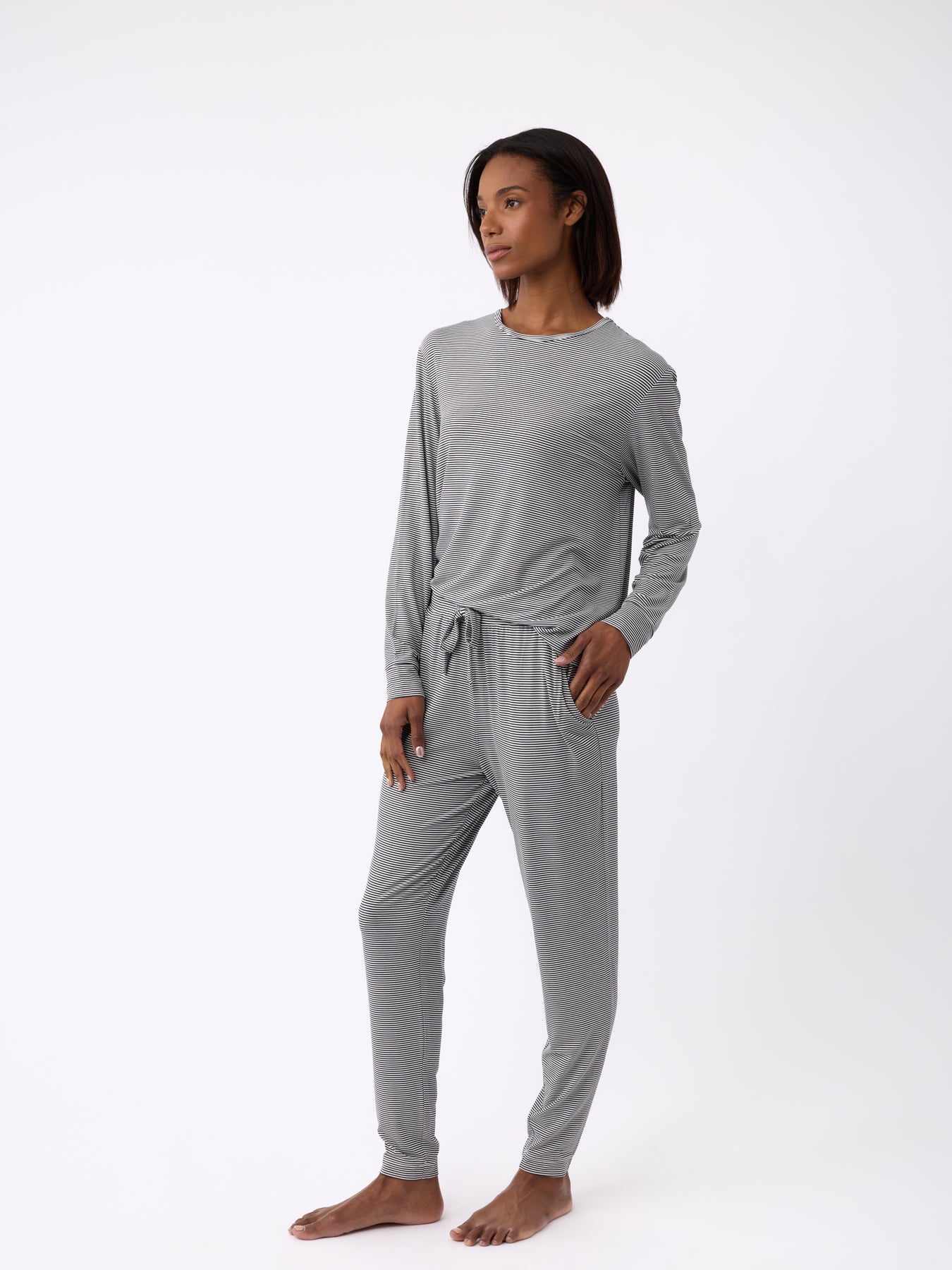 A person in Cozy Earth's Women's Bamboo Stretch-Knit Tapered Pant, gray and soft-looking with pockets, stands barefoot against a white background. They have one hand in a pocket and are gazing to the side. 