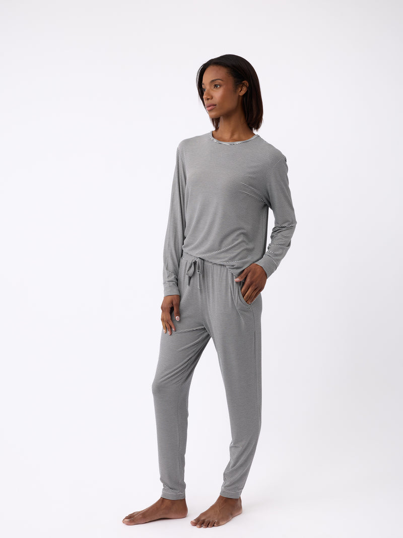 A person in Cozy Earth's Women's Bamboo Stretch-Knit Tapered Pant, gray and soft-looking with pockets, stands barefoot against a white background. They have one hand in a pocket and are gazing to the side.