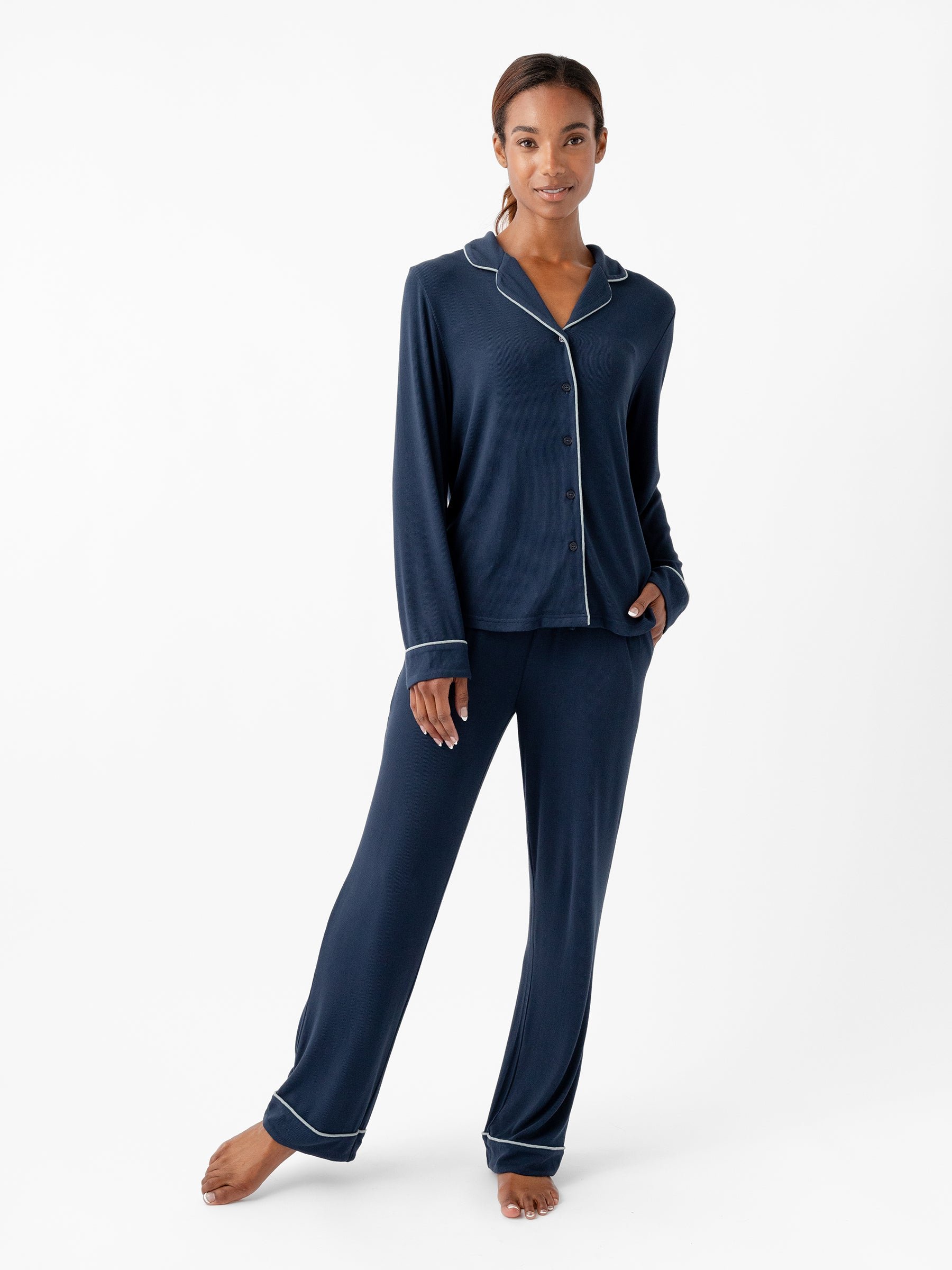 A woman stands against a plain white background, wearing the Cozy Earth Women's Bamboo Rib Knit Classic Long Sleeve Pajama Top in navy blue with white piping. The top features a collar and buttons down the front. She has one hand in her pocket and smiles softly at the camera. |Color:Eclipse