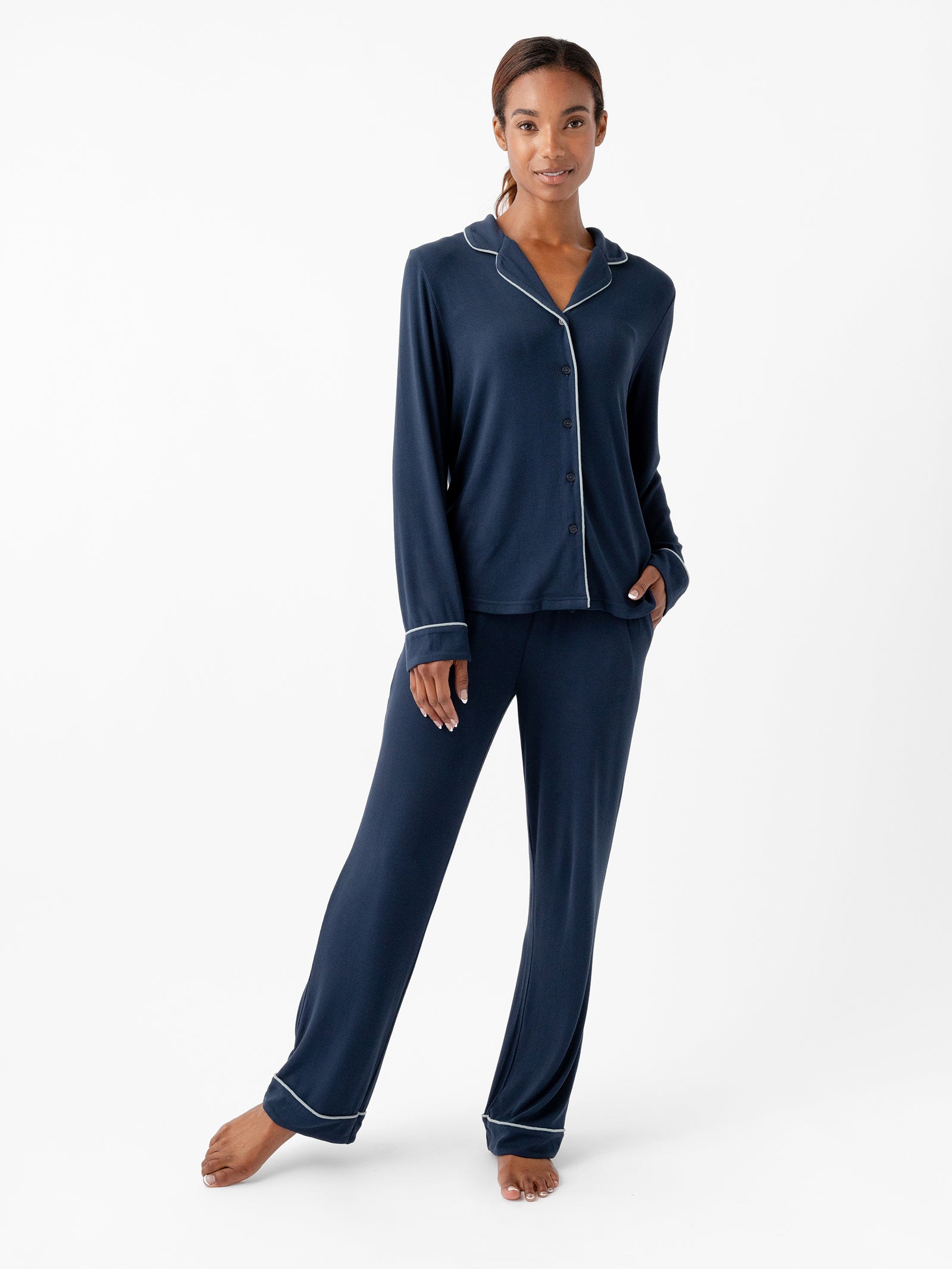 A person stands barefoot in a relaxed pose, dressed in the Cozy Earth Women's Bamboo Rib Knit Classic Pajama Pant set, styled in navy blue with white piping. This elegant pajama set features a button-up long-sleeve top with a collar and matching loose-fitting pants. The background is plain and white. 