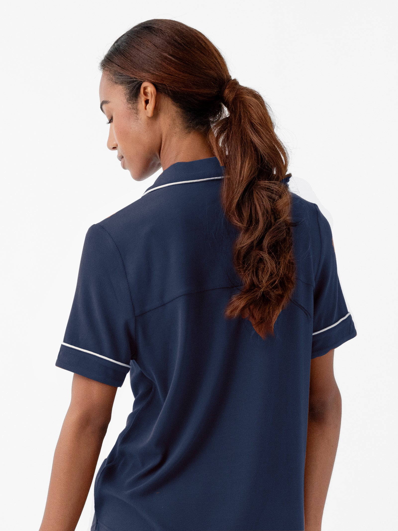 A person with brown hair tied in a ponytail is facing away from the camera, wearing a Cozy Earth Women's Bamboo Rib Knit Classic Short Sleeve Pajama Top in dark blue with white trim on the collar and sleeves. The background is plain white. 