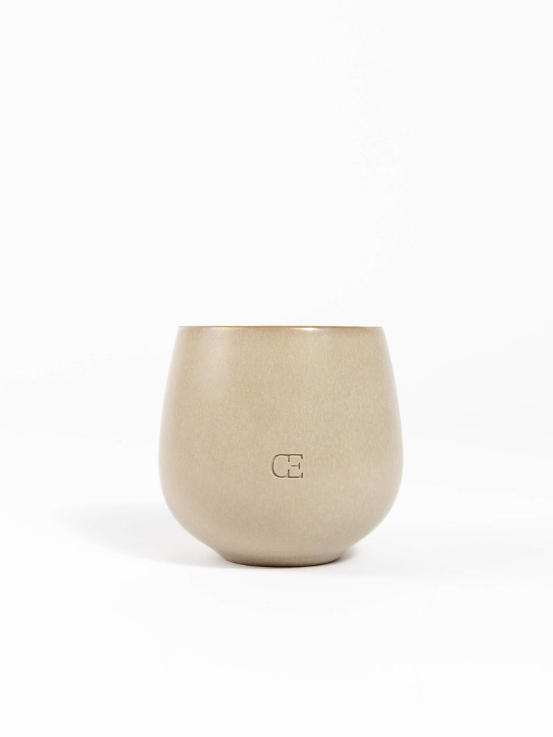 A beige, round ceramic Candle EMBER by Cozy Earth with a matte finish and "CE" embossed on the front, shown against a plain white background.