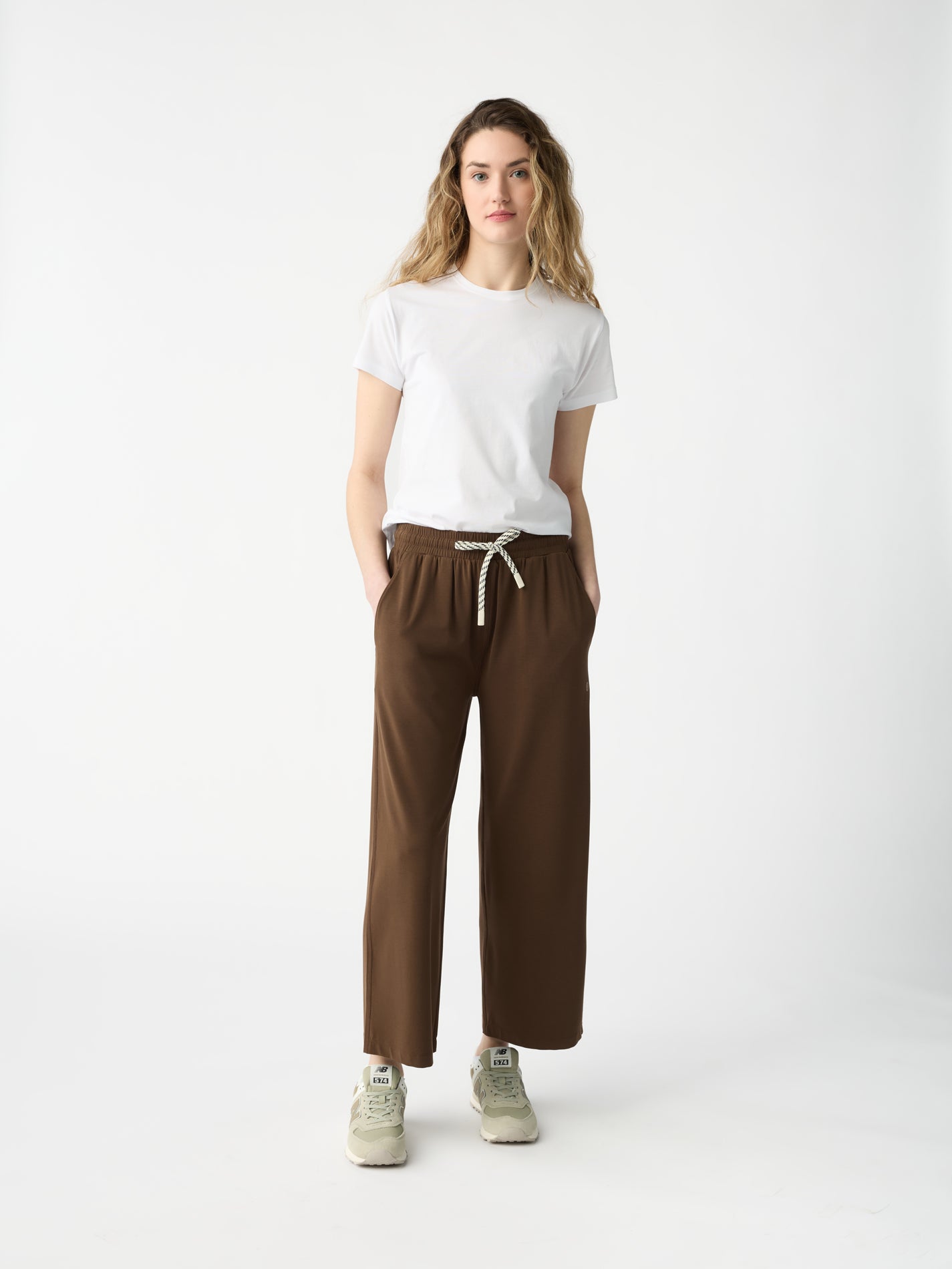 A person with long wavy hair stands against a white background, wearing a white T-shirt, Cozy Earth's Women's Studio Cropped Wide Leg Pant in brown with a drawstring, and beige sneakers. Their hands are in their pockets. 