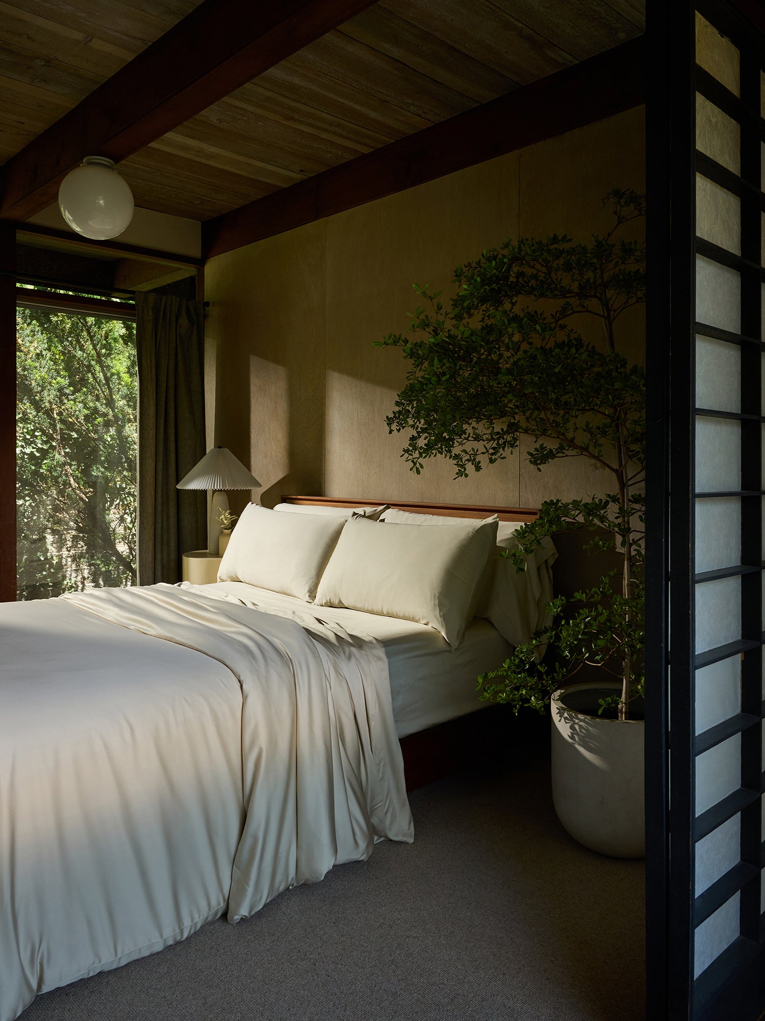 A cozy bedroom featuring a neatly made bed adorned with the Bamboo Duvet Cover by Cozy Earth. A floor lamp and a large potted plant are positioned near the bed, adding a touch of nature to the room. Natural light shines through a window with curtains, highlighting the earthy tones and wooden accents. 