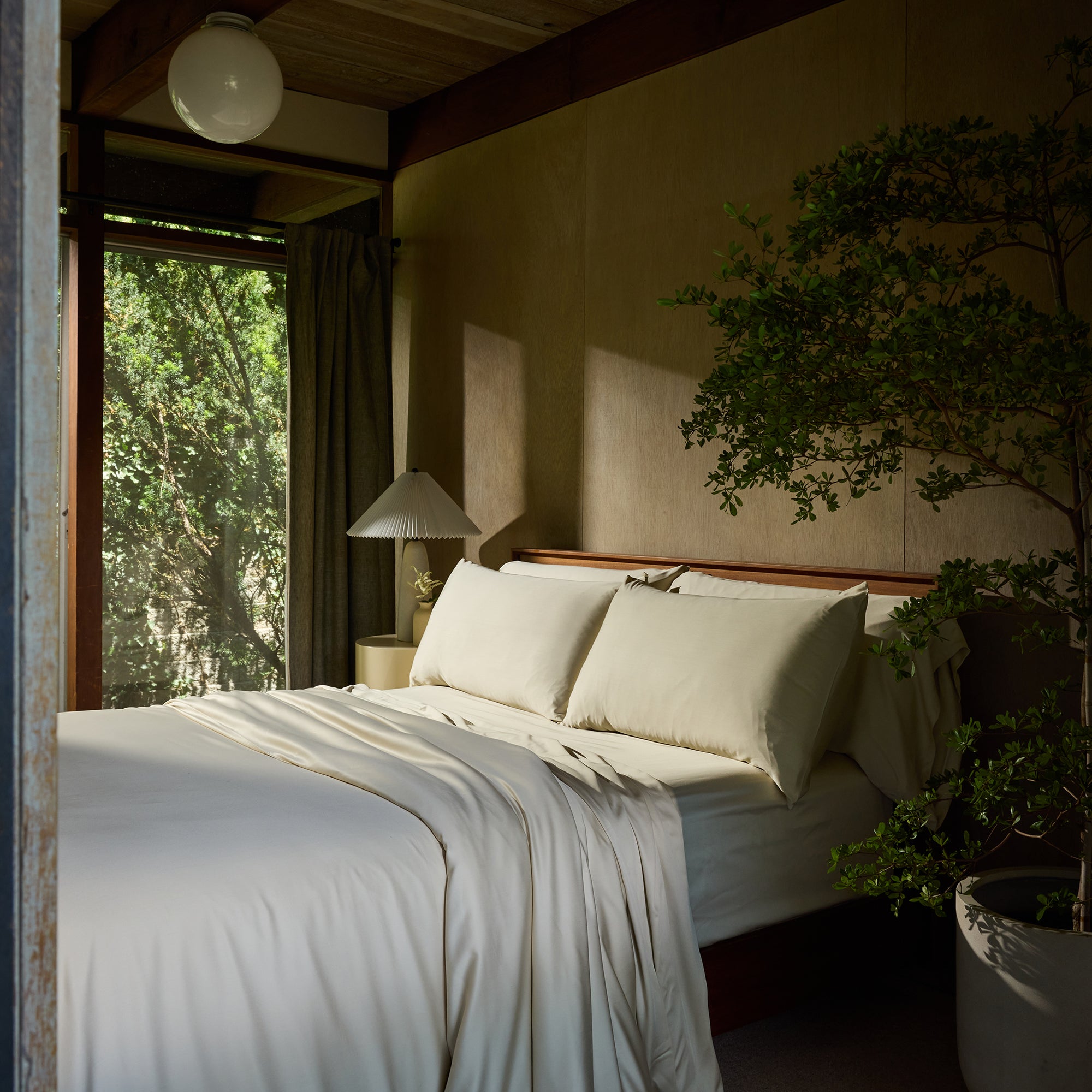 A cozy bedroom featuring a neatly made bed with Bamboo Sheet Set from Cozy Earth is adorned with a modern table lamp on a nightstand. The room includes large windows partially covered by dark curtains, allowing natural light and a view of lush greenery outside. A potted tree stands beside the bed. 