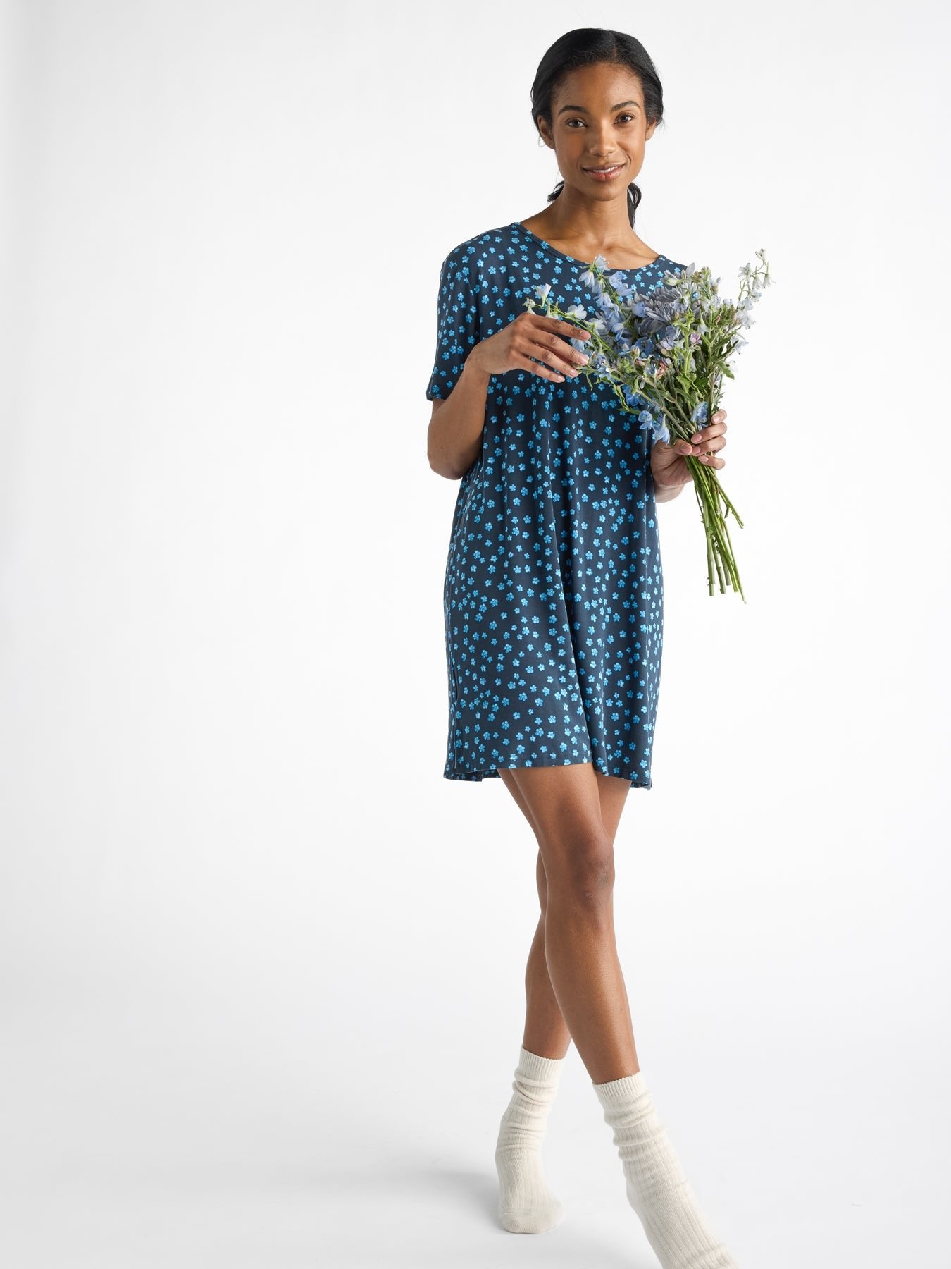 A person in a Women's Bamboo Stretch-Knit Sleep Dress by Cozy Earth holds a bouquet, walking barefoot in cream socks against a white background. 