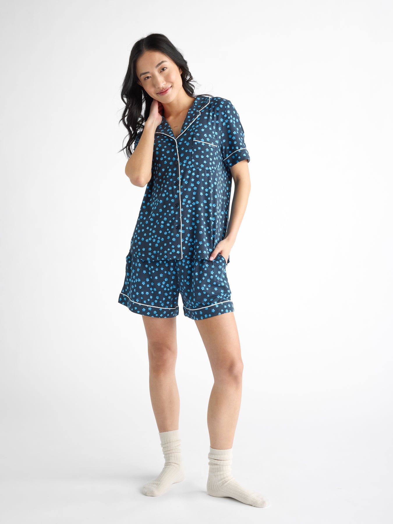 A person wearing a blue polka dot, Women's Bamboo Stretch-Knit Short Sleeve Pajama Top from HIDE with white trim smiles with one hand on their chin. They also have on white socks against a plain white background. 