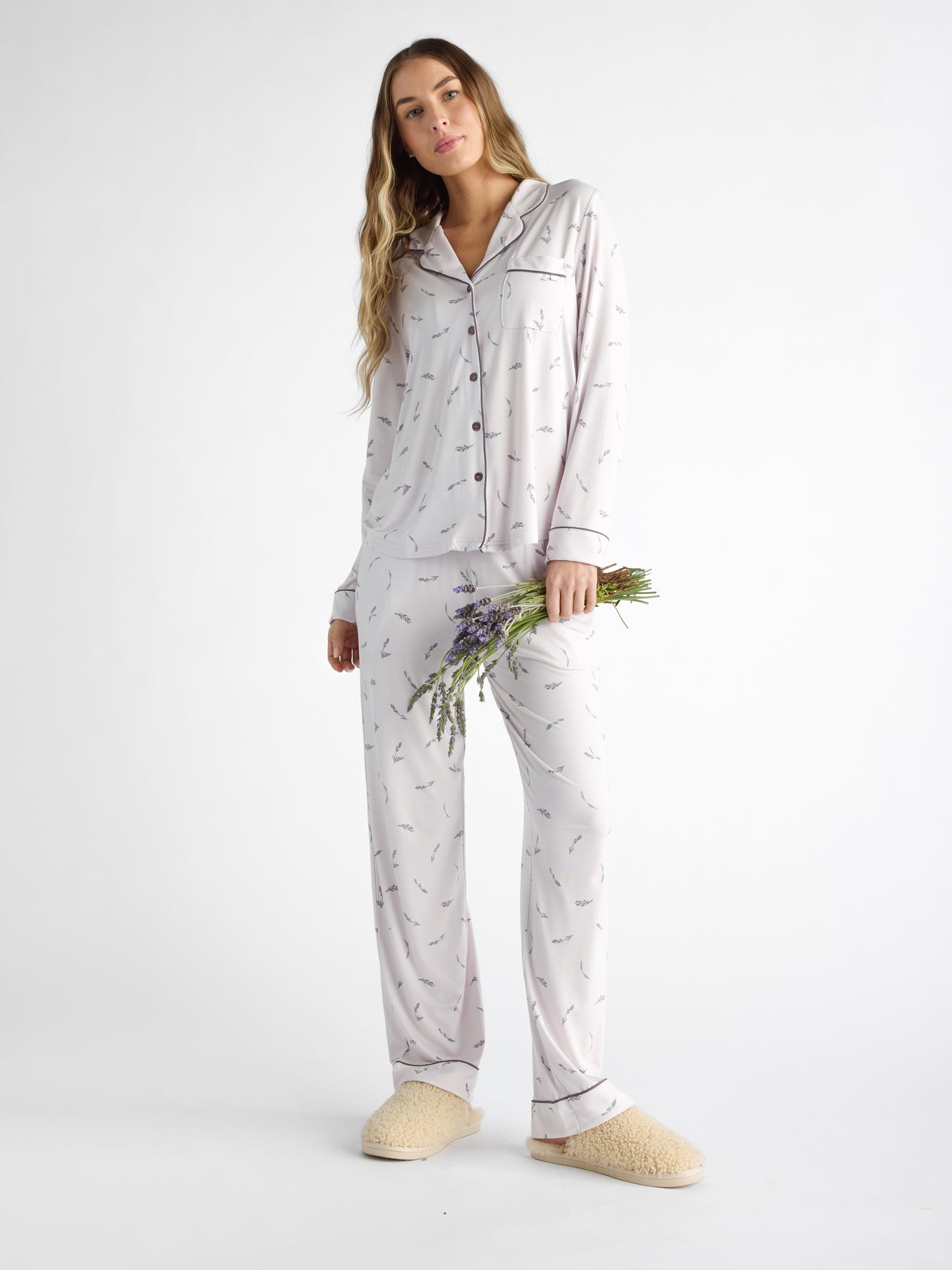 Against a plain background, a person wears Cozy earth Women's Bamboo Stretch-Knit Pajama Pant in a light color with a subtle pattern, paired with beige slippers. They hold a small lavender bouquet as their long, wavy hair cascades down. 