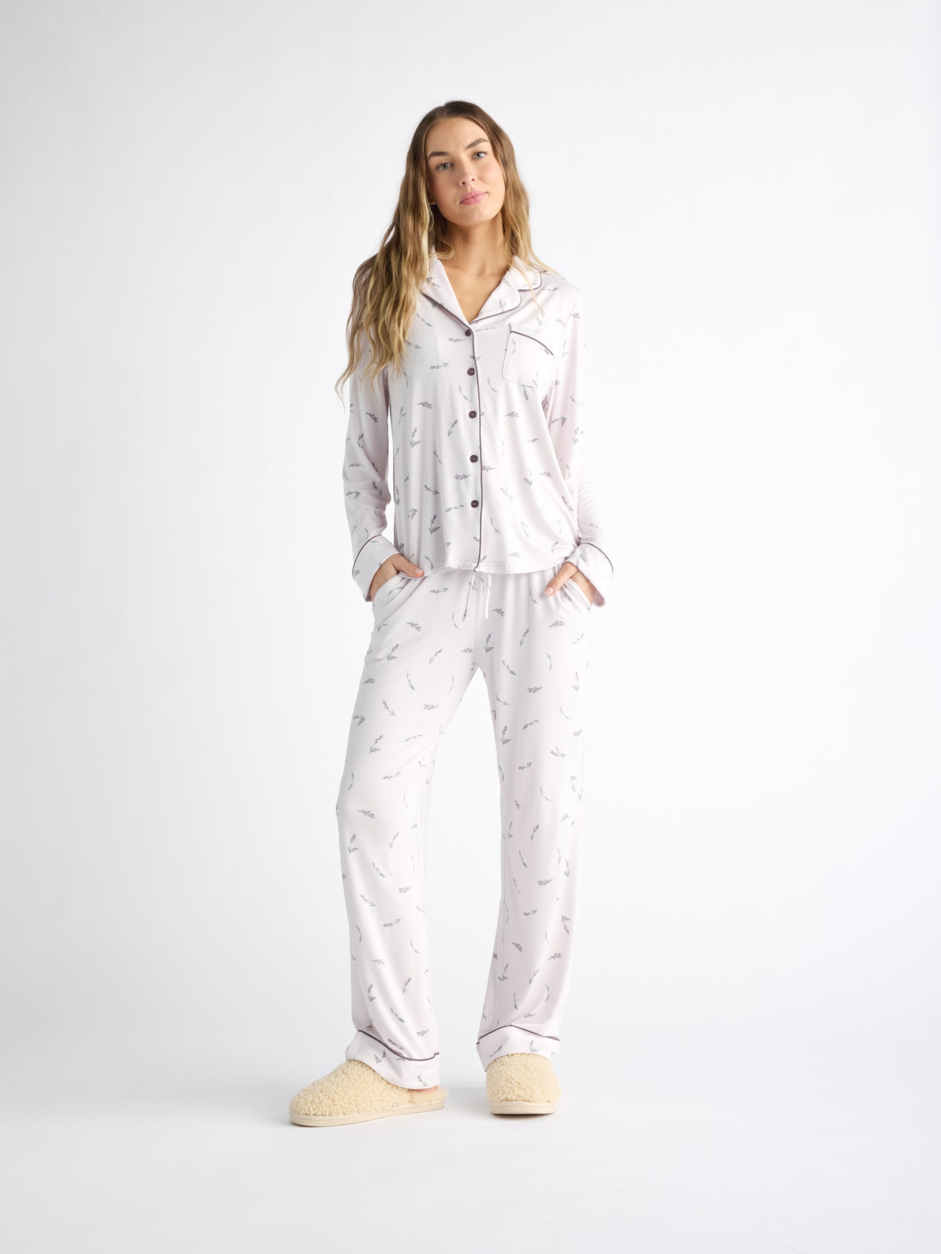 A woman stands against a plain white background wearing the Cozy Earth Women's Bamboo Stretch-Knit Long Sleeve Pajama Set and fluffy slippers. She has long hair and her hands are tucked into the pockets of her pajama pants. |Color:French Lavender