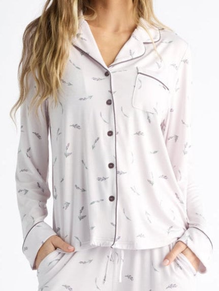 A woman wearing the Women's Bamboo Stretch-Knit Long Sleeve Pajama Top in a light color with patterns and beige slippers stands in a relaxed pose. She has long hair, set against a plain white background.