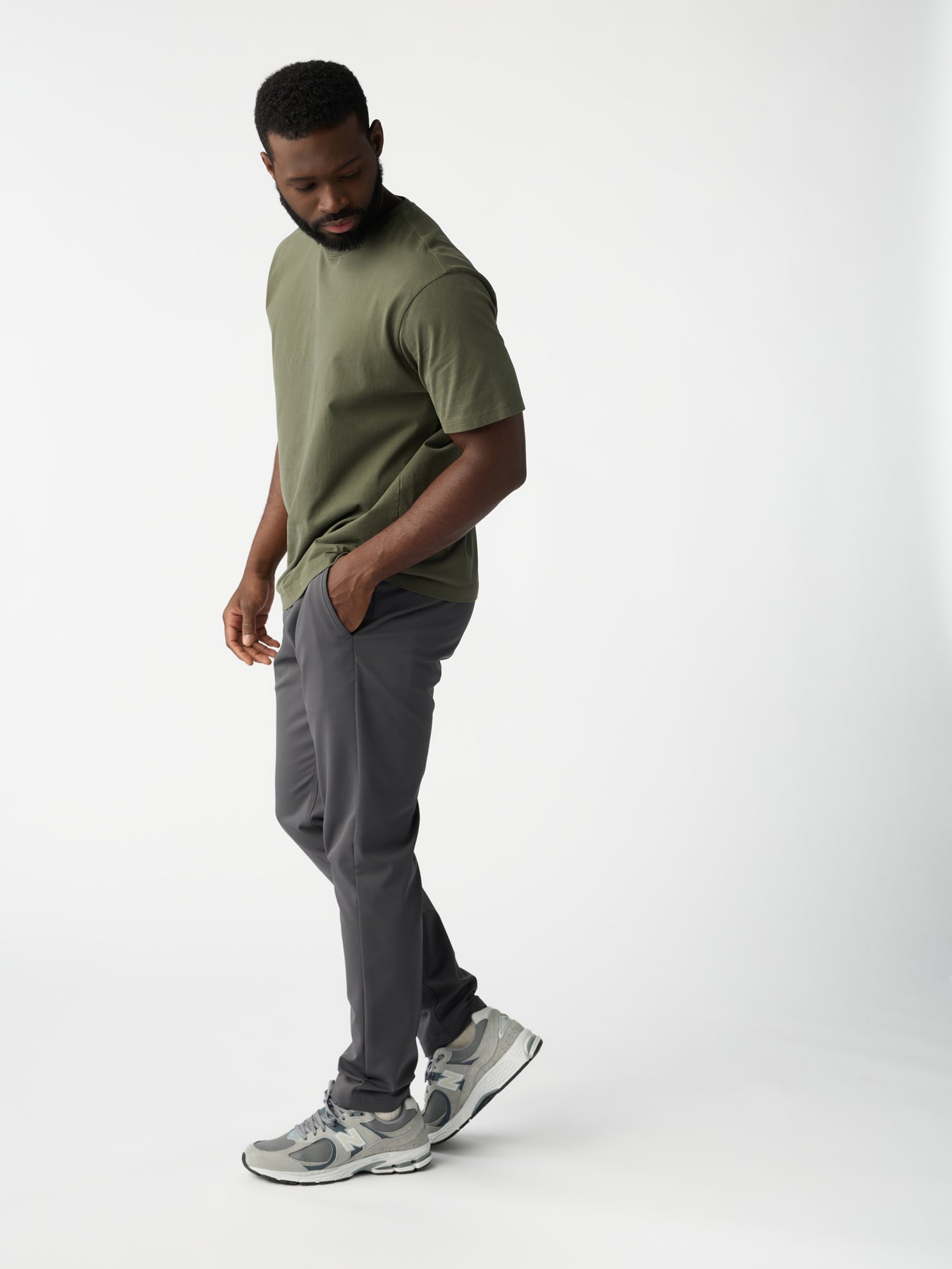 A man sporting a Cozy Earth olive green T-shirt, paired with Men's Everywhere Pant in gray and gray sneakers, stands against a white background. He's looking down, one hand in his pocket, exuding a relaxed demeanor. 