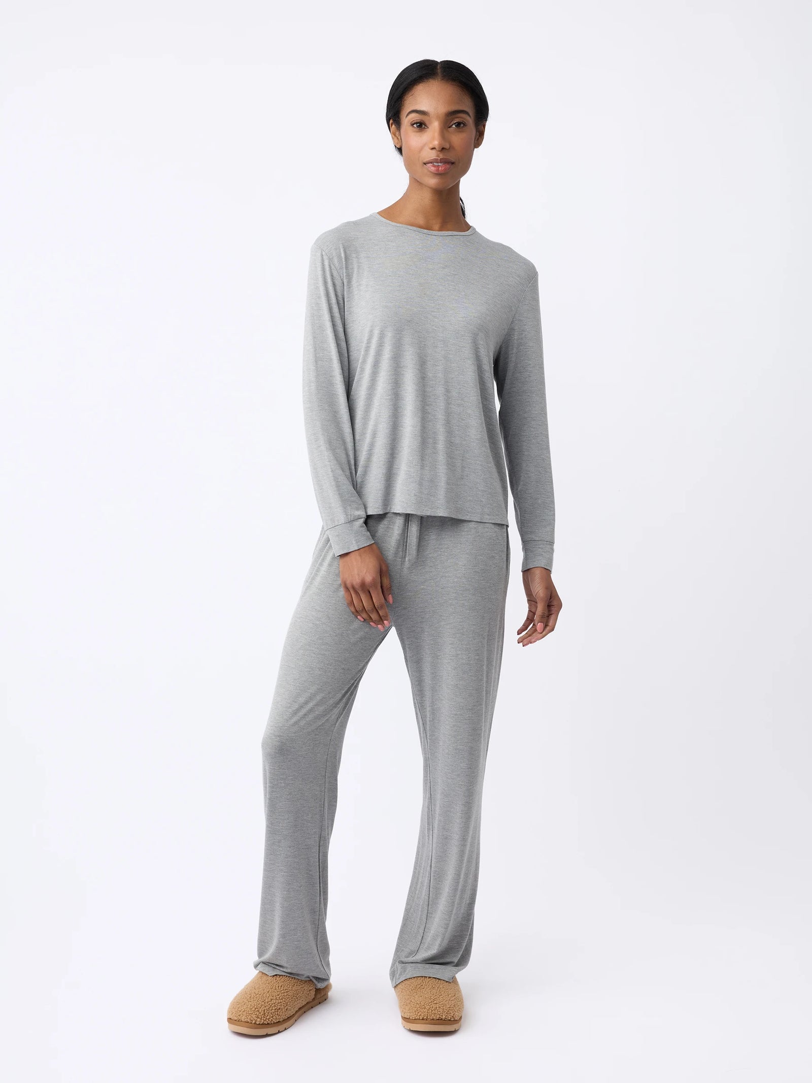 A woman stands against a plain white background, wearing Cozy Earth's Women's Stretch Knit Bamboo Long Sleeve Lounge Tee in light gray with matching loose-fitting pants and tan slippers. They have a calm expression. 