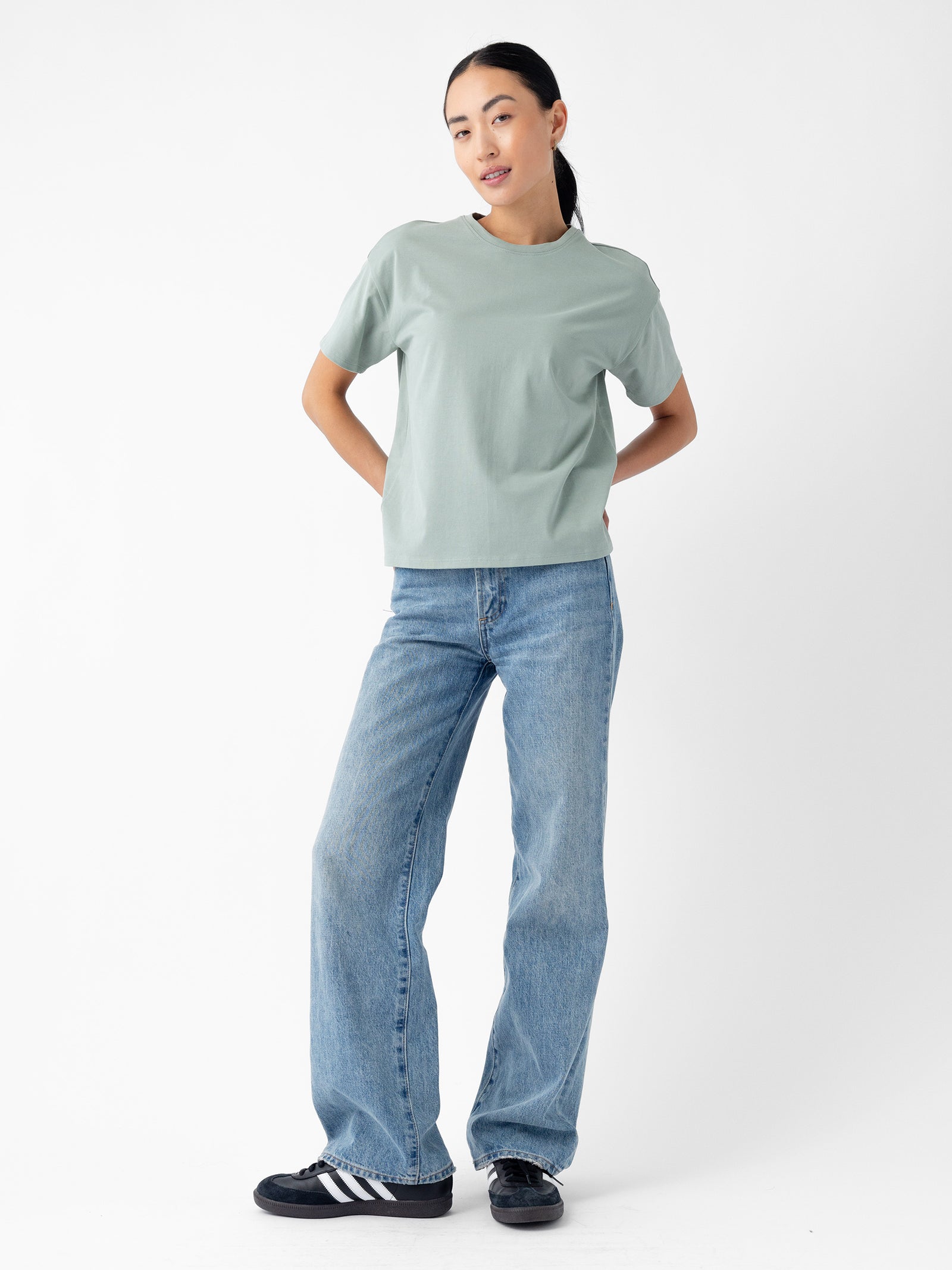 A person stands against a plain white background wearing the Women's All Day Tee in light green from Cozy Earth, blue jeans, and black sneakers with white stripes. They have long dark hair tied back and are posing with hands on hips, looking towards the camera. 