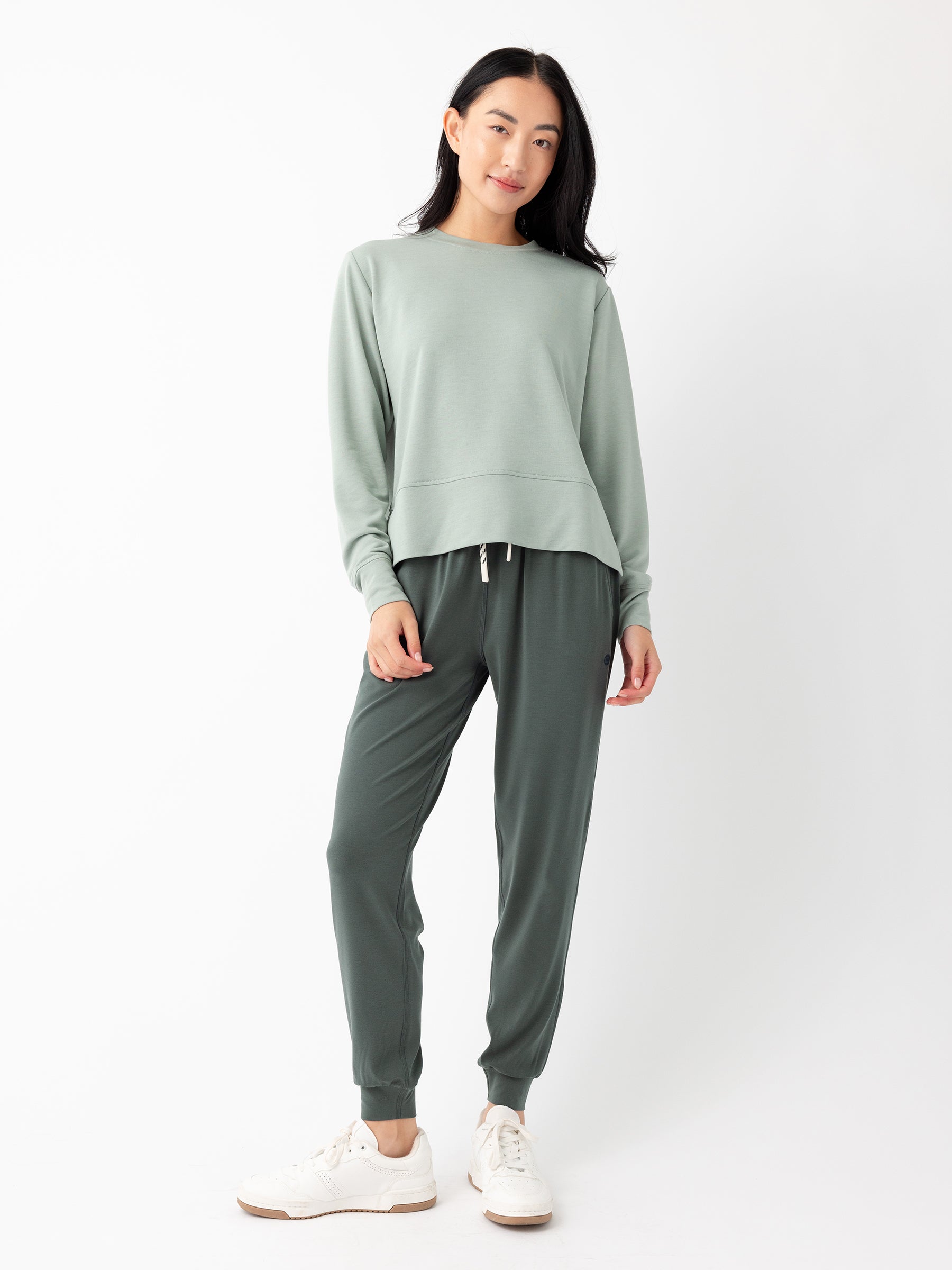 A woman with long dark hair stands against a white background, smiling slightly. She is wearing Cozy Earth's Women's StudioLite Crewneck in light green and dark green jogger-style pants, along with white sneakers. |Color:Haze