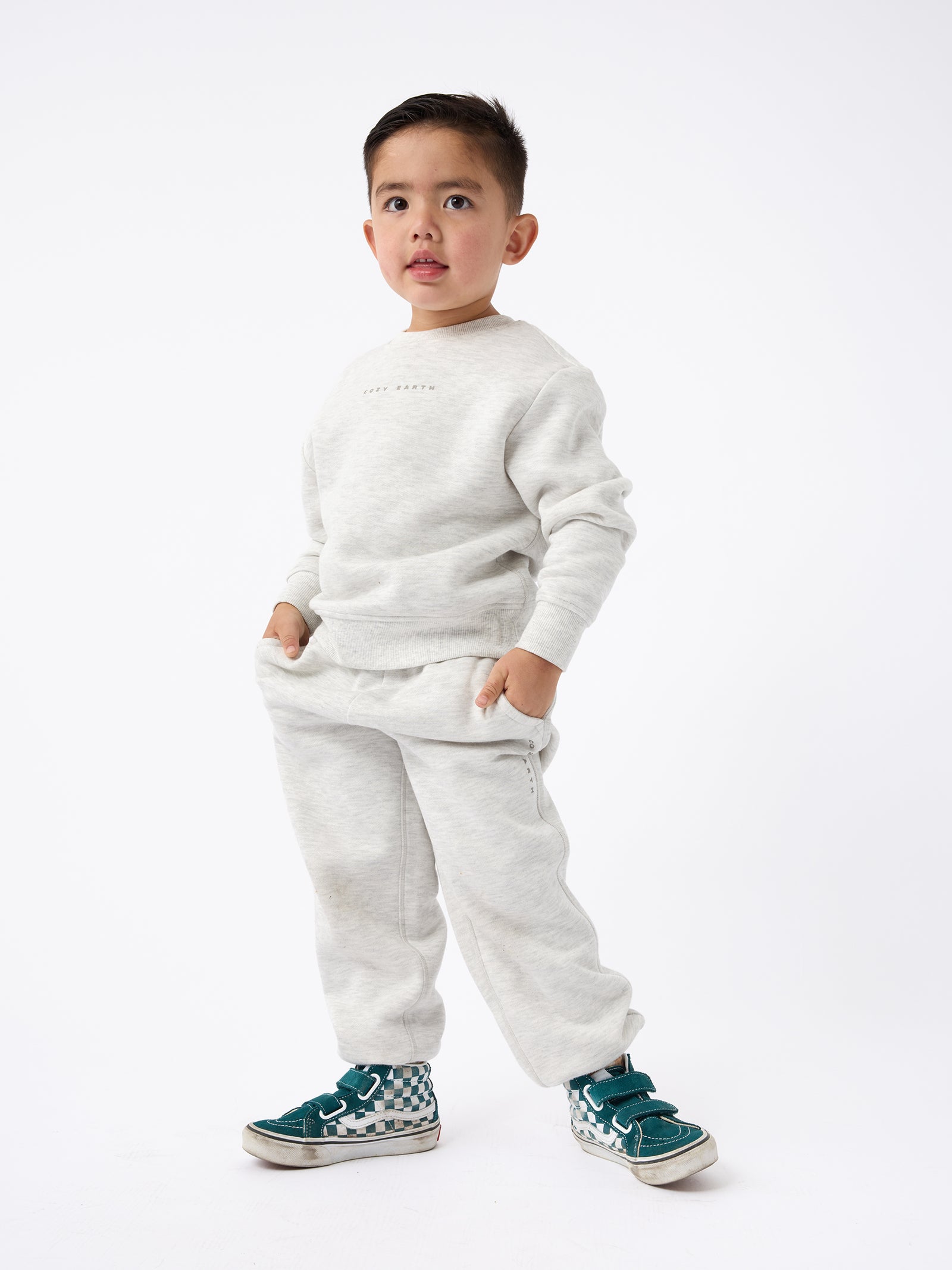 A young child stands confidently against a plain background, dressed in the Kid's CityScape Crewneck by Cozy Earth and sporting green sneakers. With hands tucked into pockets, the child strikes a relaxed pose. The outfit looks comfortable and casual. 