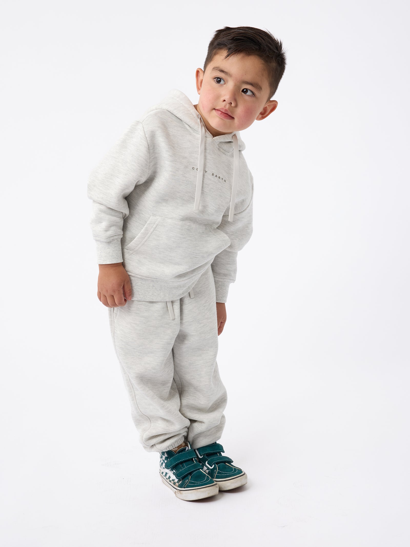 A young boy wearing the Cozy Earth's Kid's CityScape Hoodie in light gray, along with matching sweatpants, leans to the side with a curious expression. He has short hair and is sporting teal sneakers against a plain white background. 