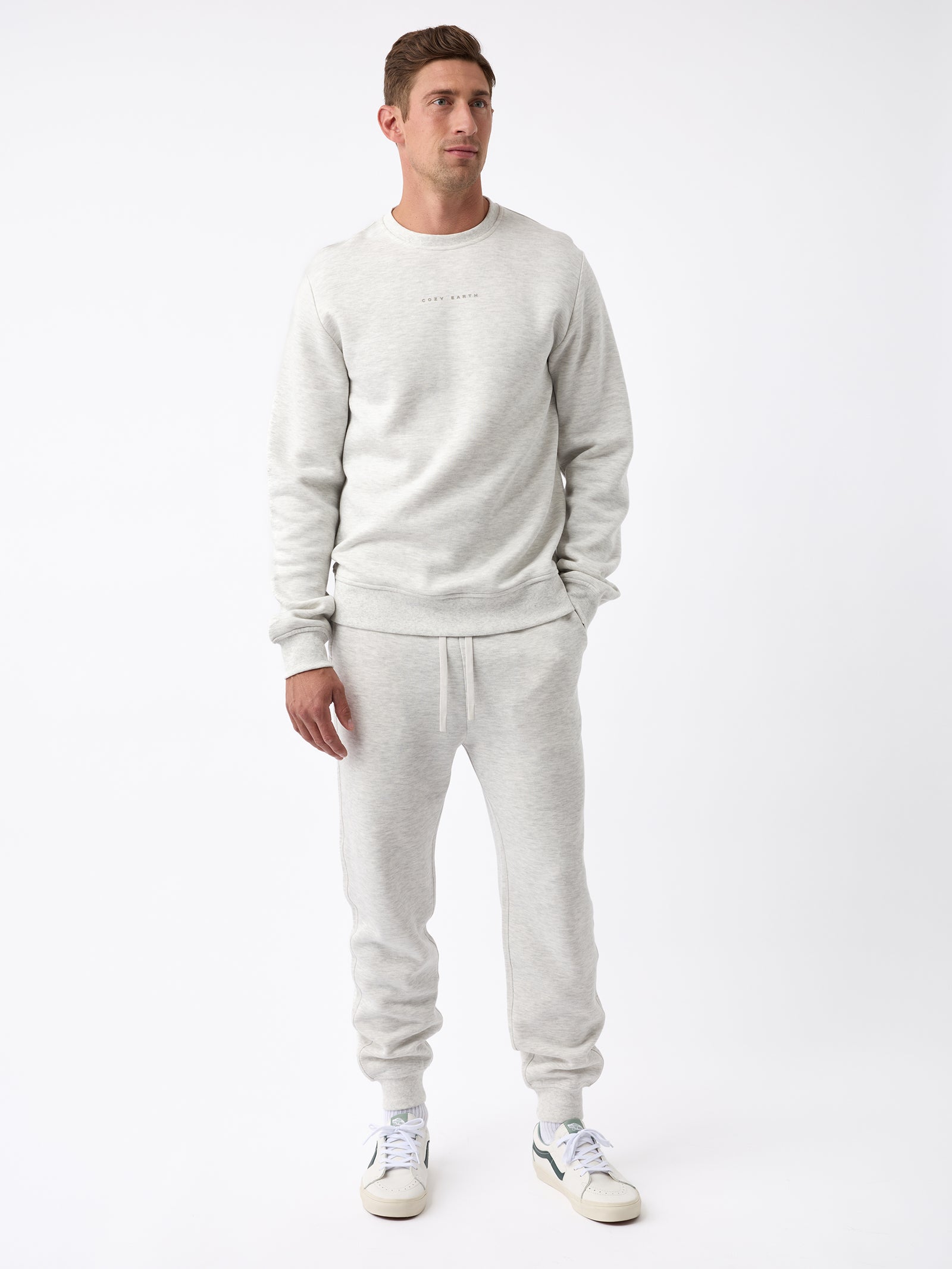 A man stands against a white background wearing a light gray Men's CityScape Jogger by Cozy Earth, paired with matching sweatpants. He is also wearing white sneakers with green accents. His hands are by his sides, and he looks slightly to his right. 