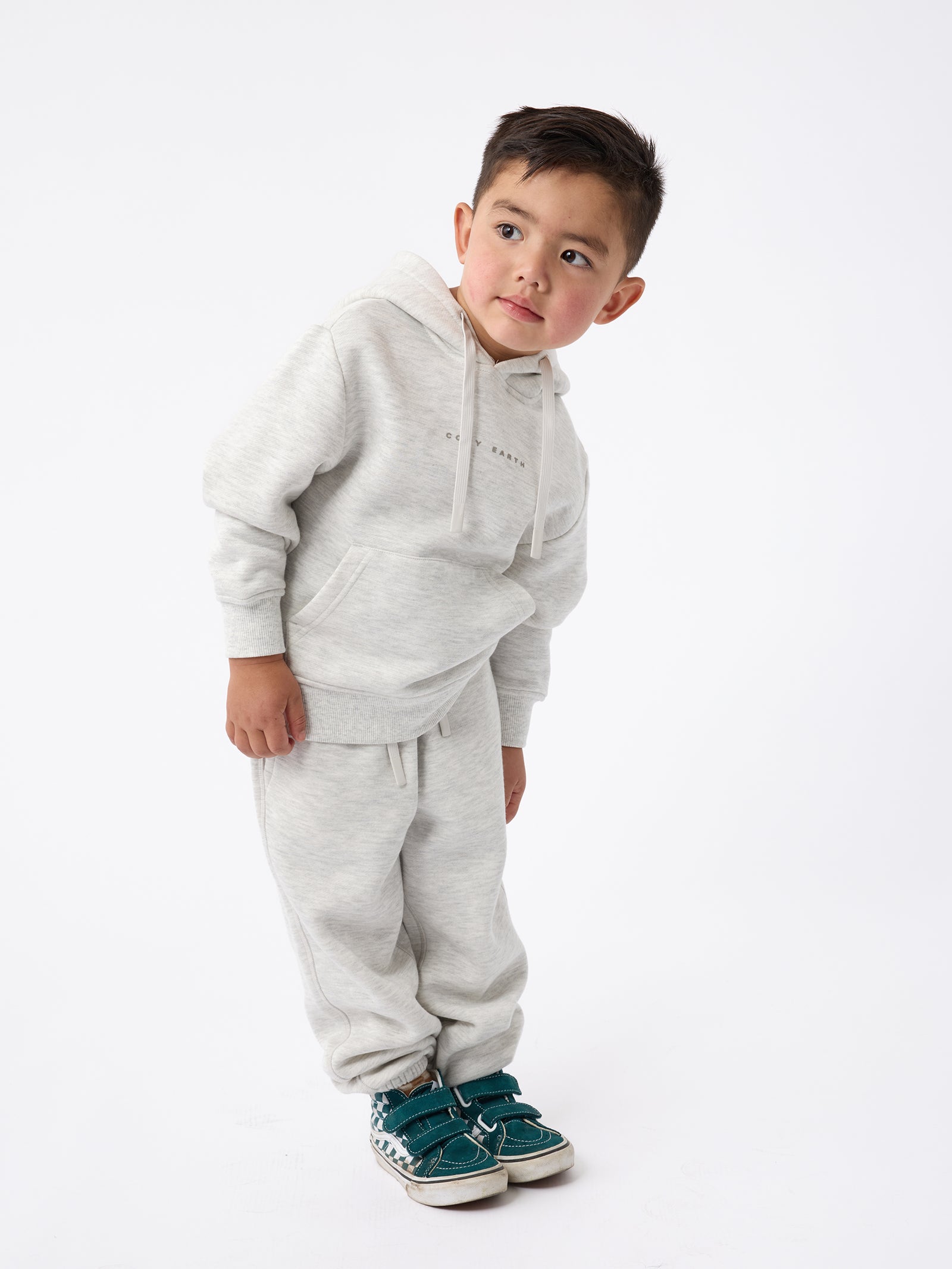 A young child dressed in Cozy Earth's Kid's CityScape Sweatpants in a light gray color stands leaning slightly to one side against a plain white background. He is wearing green sneakers and gazes thoughtfully to his right. 