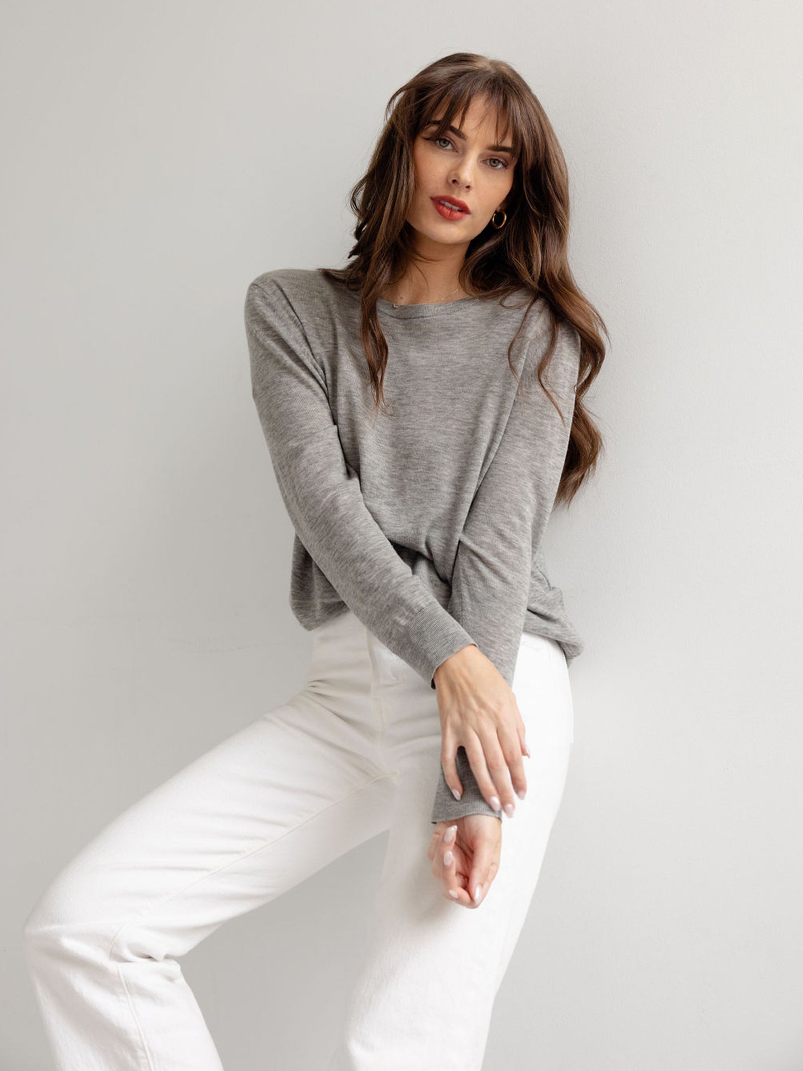 Woman leaning against wall in heather grey airknit sweater and white jeans with white background 