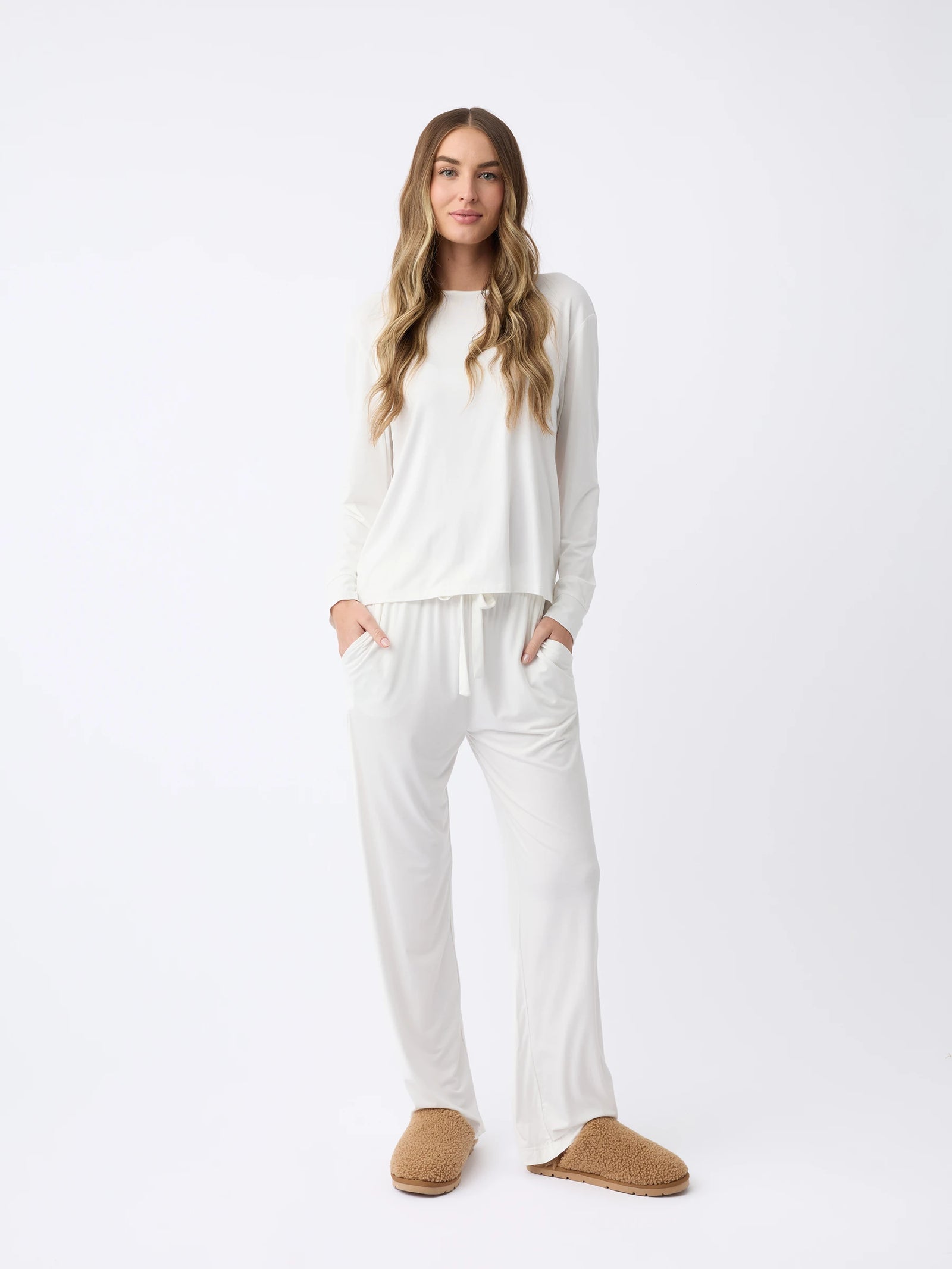 A woman models Cozy Earth's Women's Stretch Knit Bamboo Long Sleeve Lounge Tee, paired with brown slippers. Their long, wavy hair falls naturally as their hands rest in the pants pockets against a plain white backdrop. 