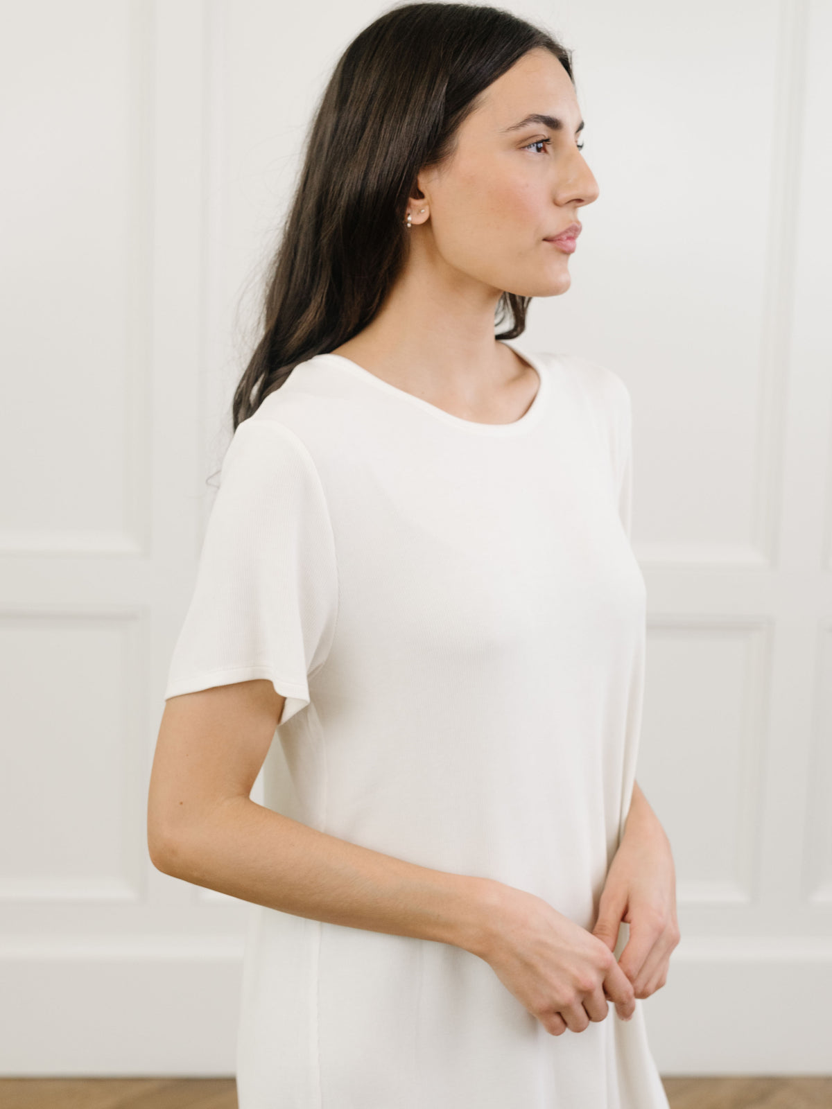 A woman standing in a white Cozy Earth bamboo rib-knit boyfriend sleep dress with short sleeves, knee-length, in a lounge or bedroom setting. 