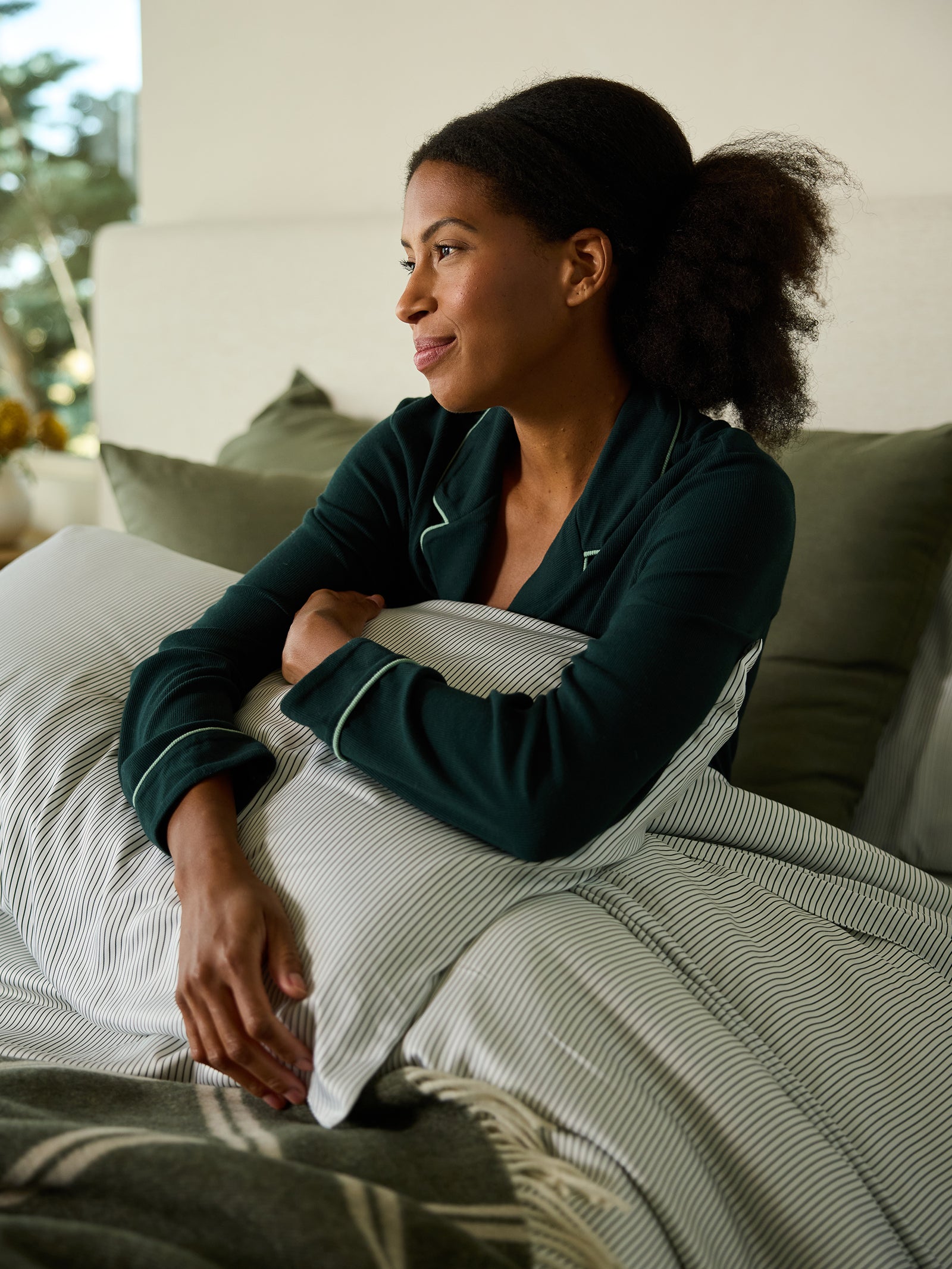 A person in dark green pajamas sits on a bed, hugging a pillow adorned with Cozy Earth's Bamboo Pillowcases. They gaze thoughtfully out the window, in a softly lit room complemented by a plant and cozy blankets in the background. 