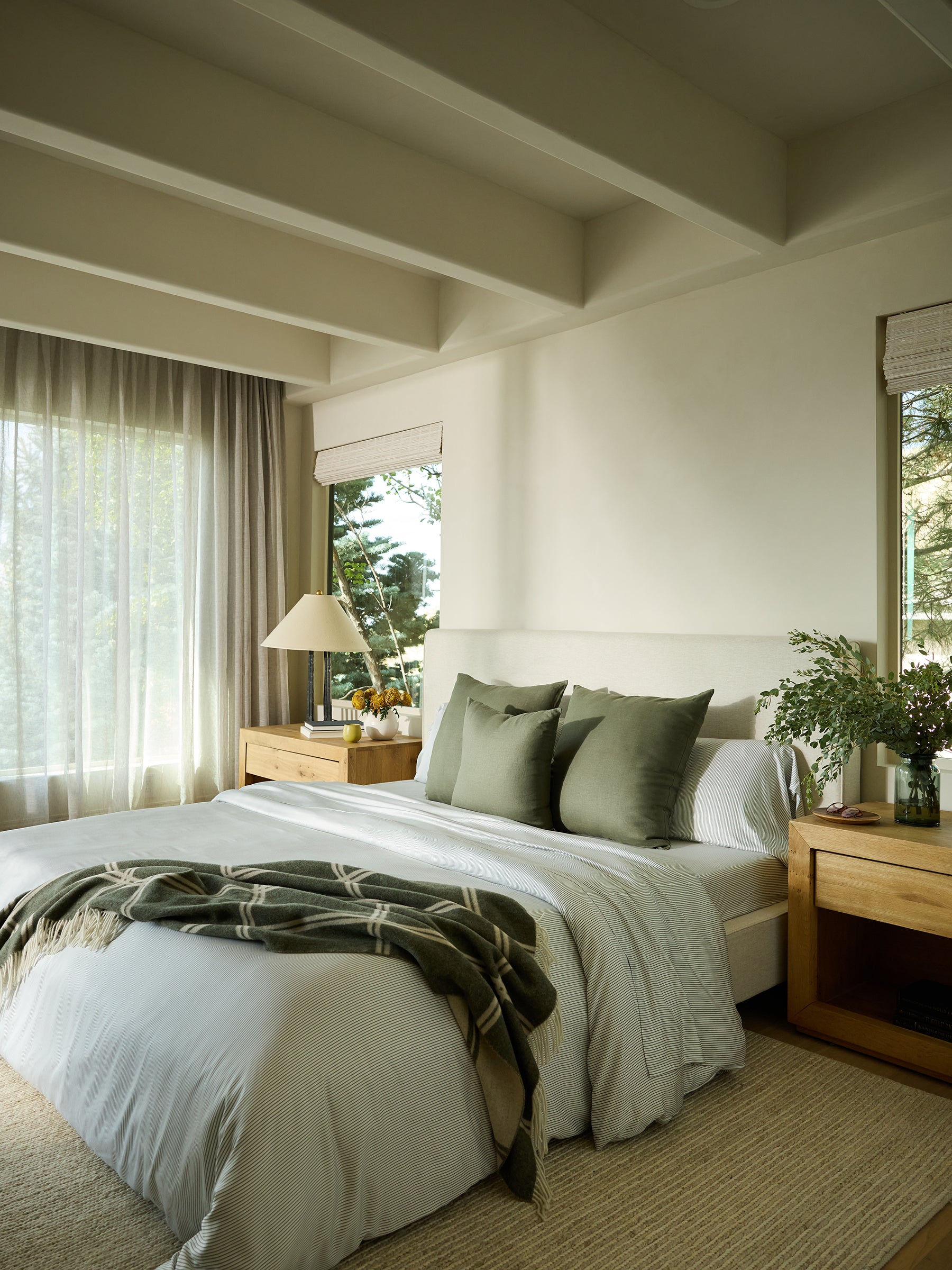 In a tranquil bedroom, a neatly made bed is adorned with Cozy Earth's Bamboo Fitted Sheet and highlighted by green pillows and a plaid throw. Gentle natural light streams through sheer curtains, casting a warm glow on the soft-colored walls and wooden furniture. Beside the bed, a table holds a lamp and a vase filled with lush greenery. 